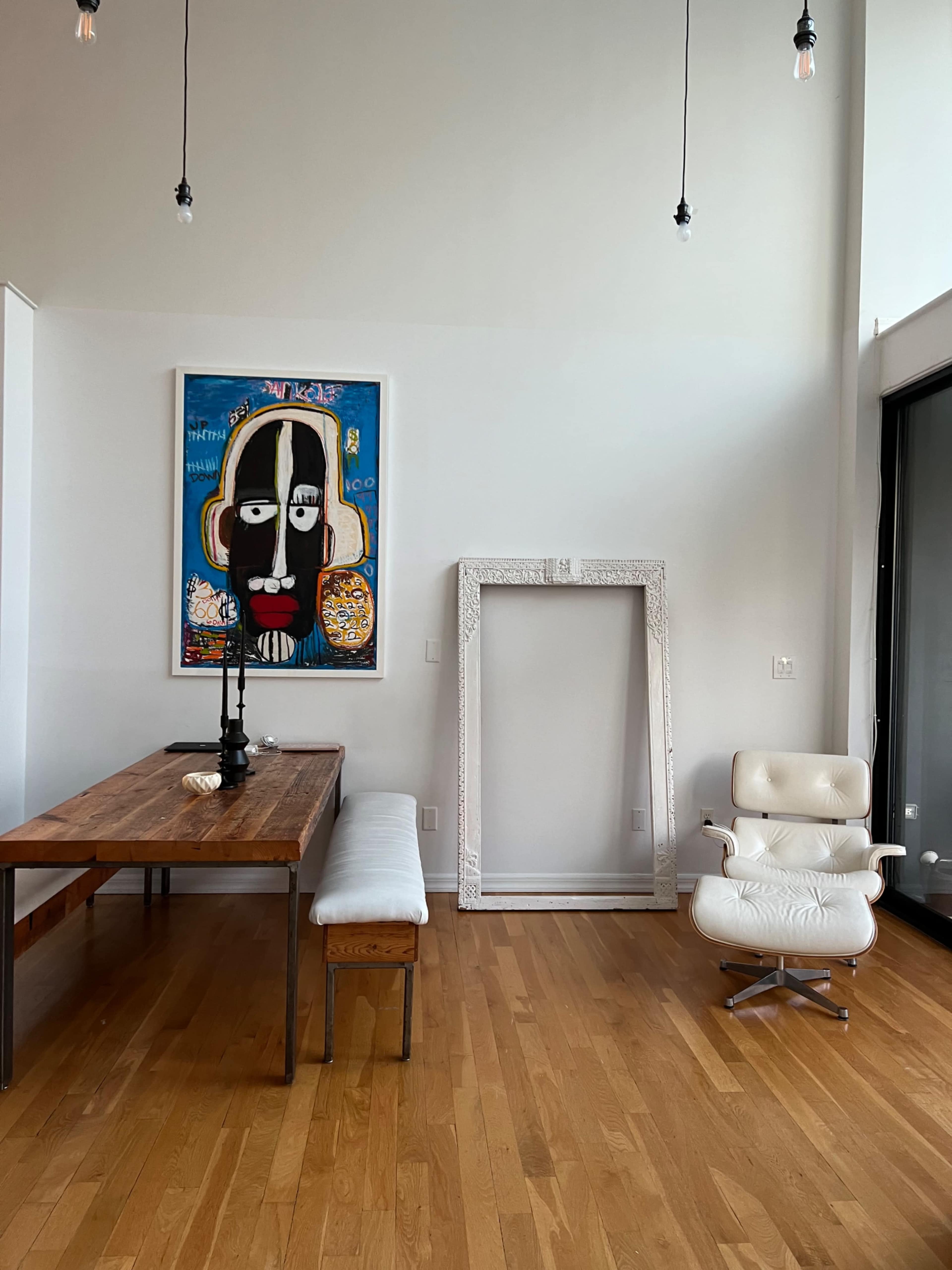 A minimalist interior with a wooden dining table, a white bench, an abstract painting on the wall, and an ornate picture frame next to a modern chair.