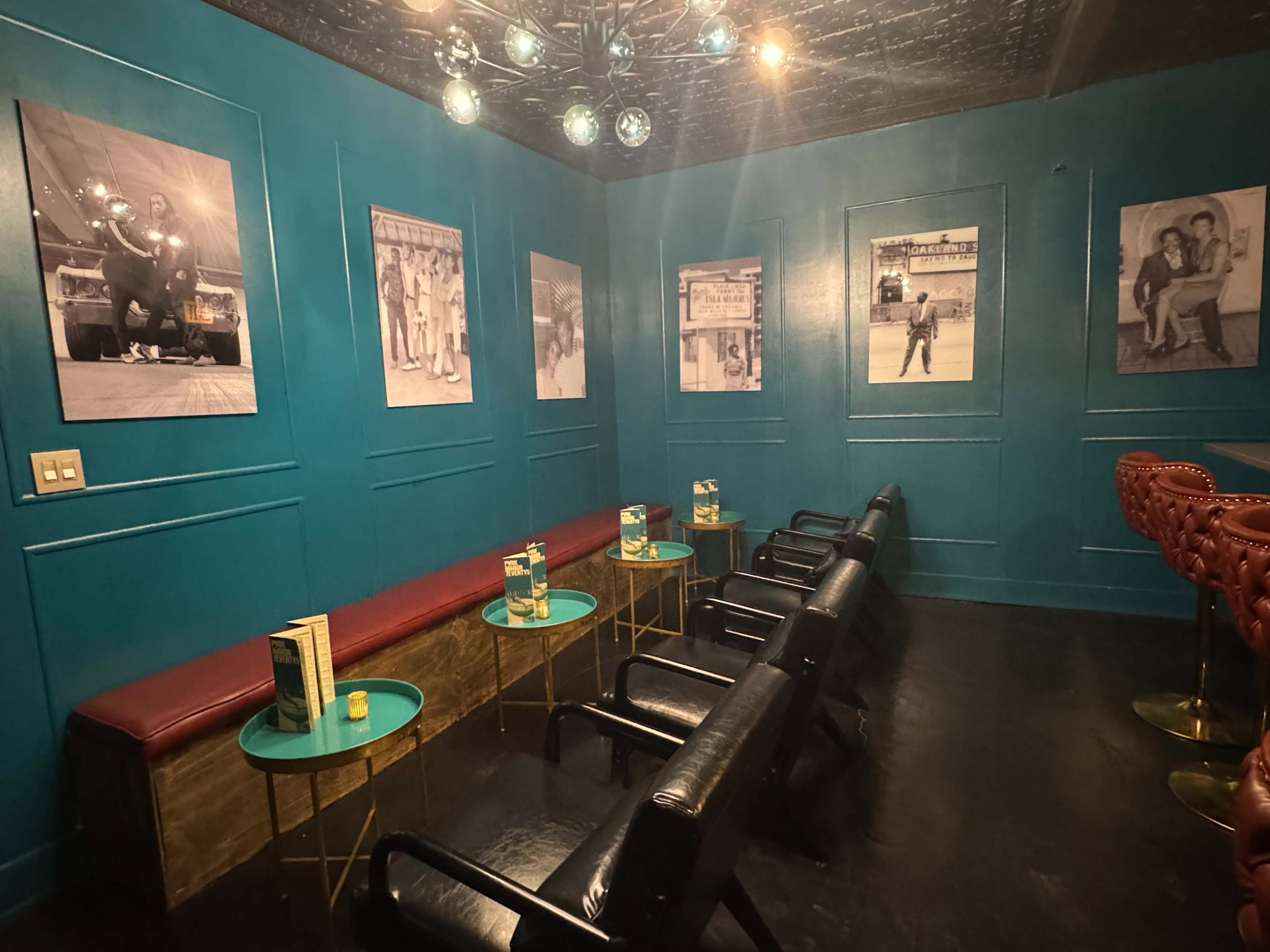 The image shows a small, teal-walled lounge area with black leather chairs arranged around gold tables, featuring vintage black-and-white photographs on the walls.