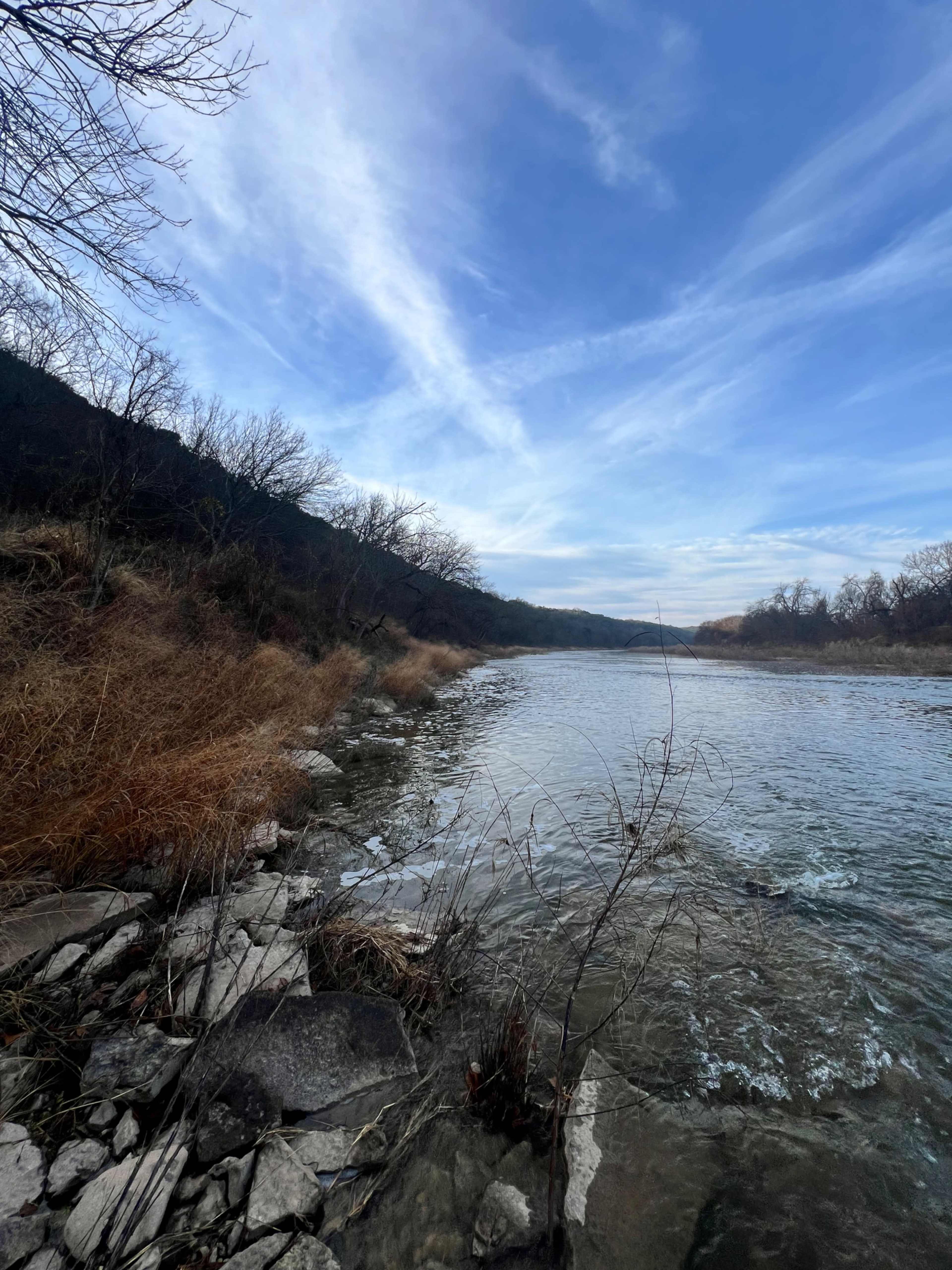 A winding river flows alongside a rocky shoreline, bordered by sparse trees and grasses under a blue sky with wispy clouds.