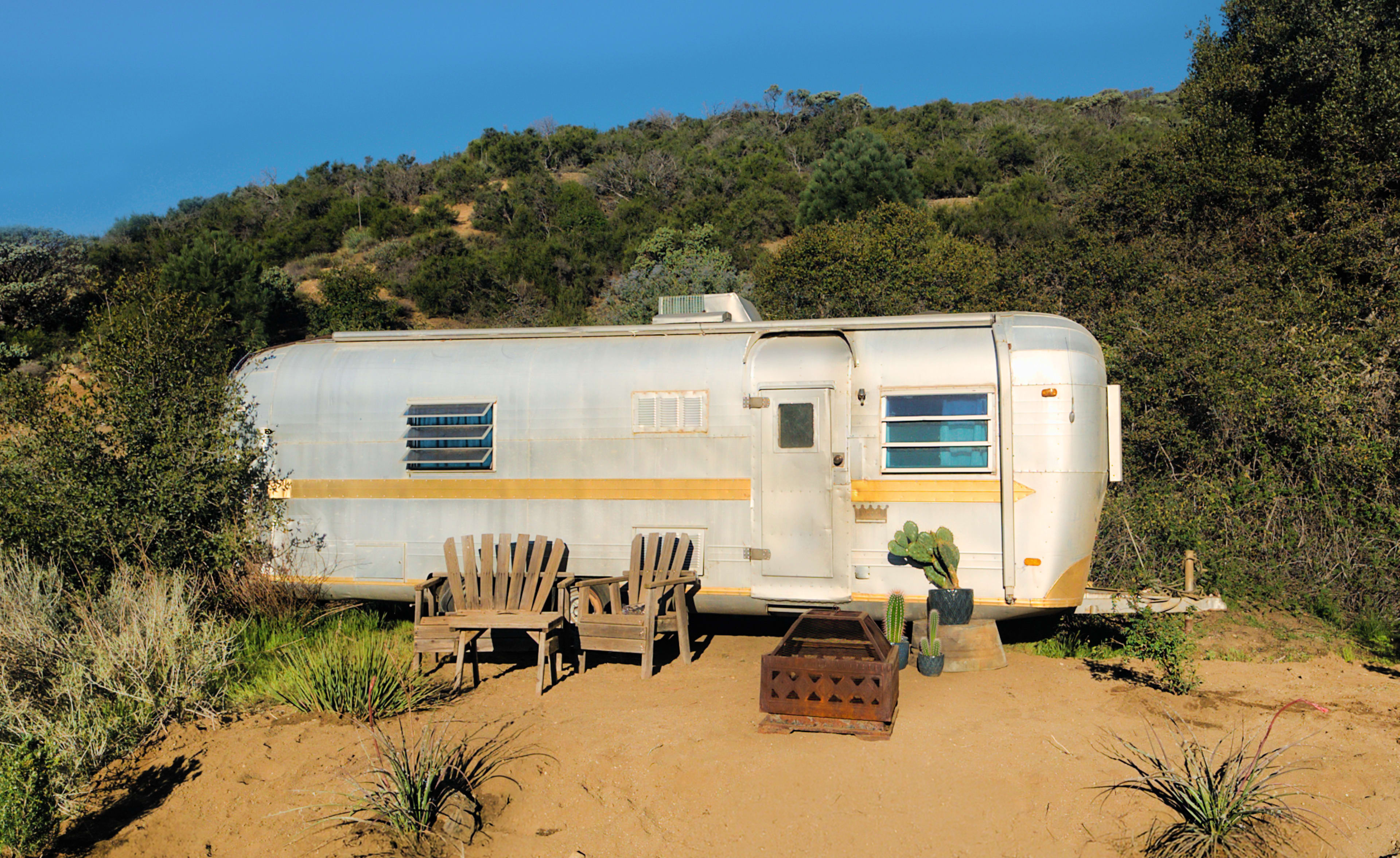 A vintage Airstream trailer is parked in a sandy area surrounded by shrubs, with two wooden chairs and a small table in front of it.