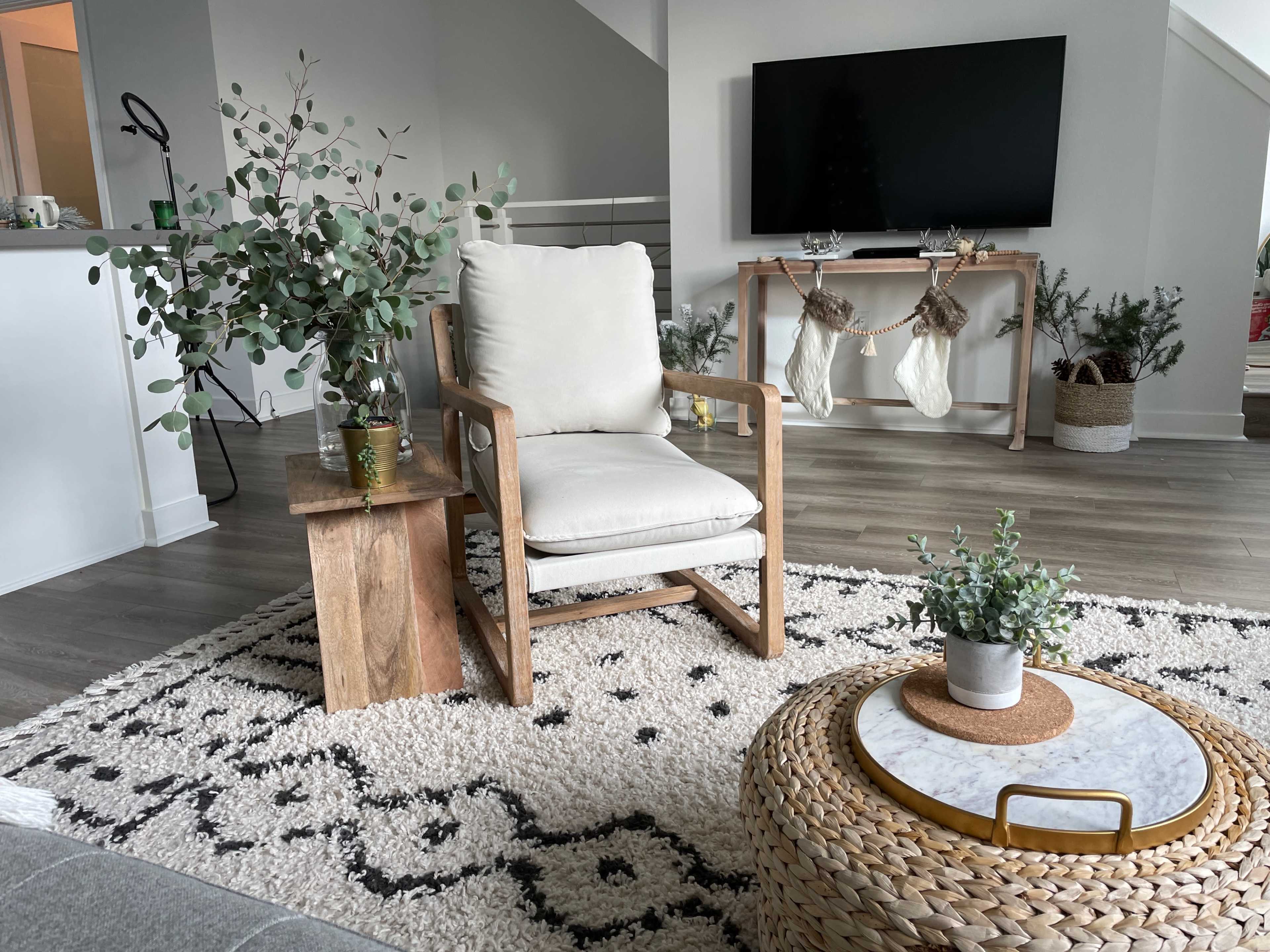 The scene features a cozy living room with a wooden chair, a small side table, a round coffee table with a marble top, and festive decorations along with plants on a textured rug.