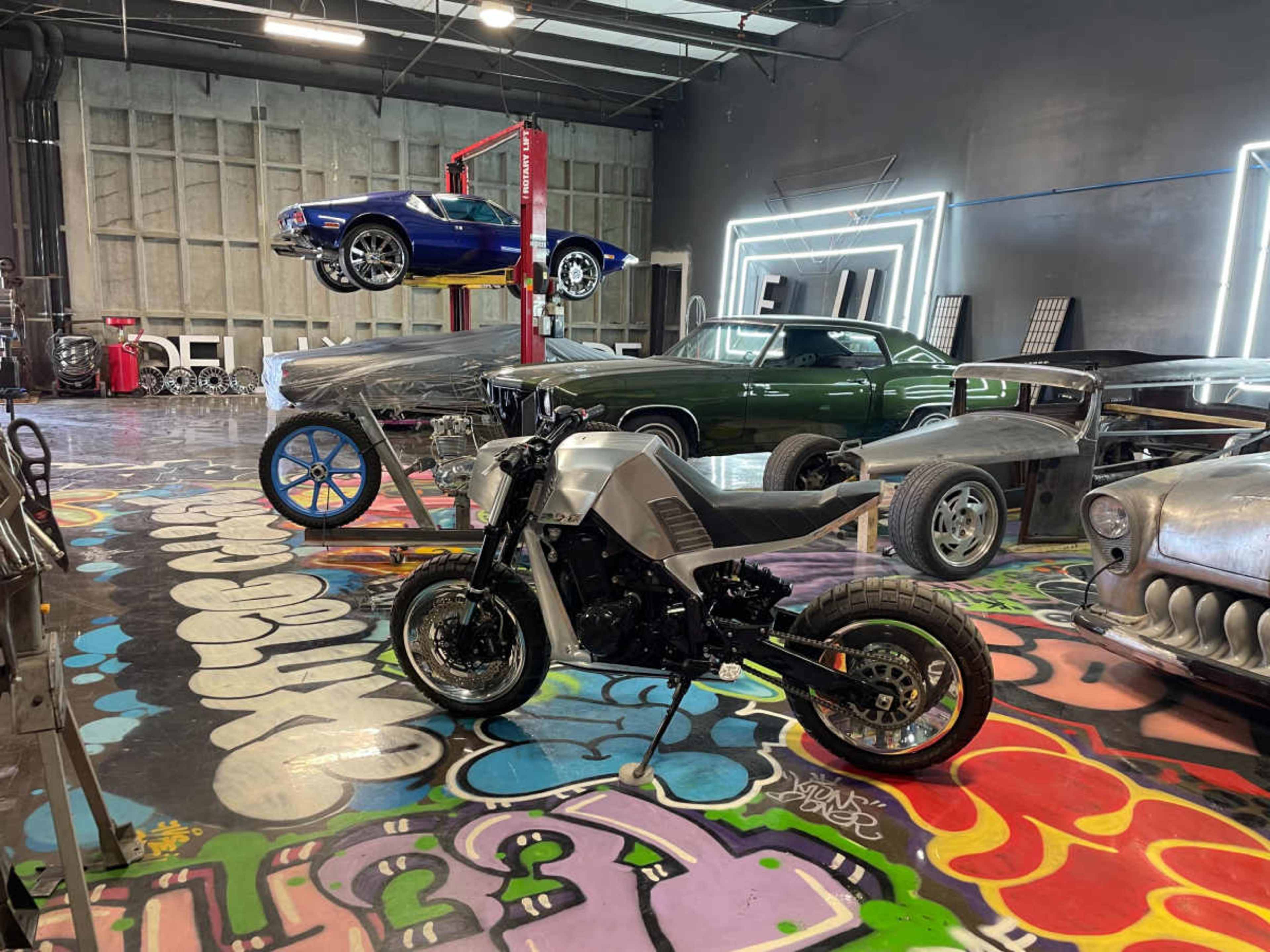 A spacious garage features a mix of classic cars and a motorcycle, with vibrant graffiti covering the floor.