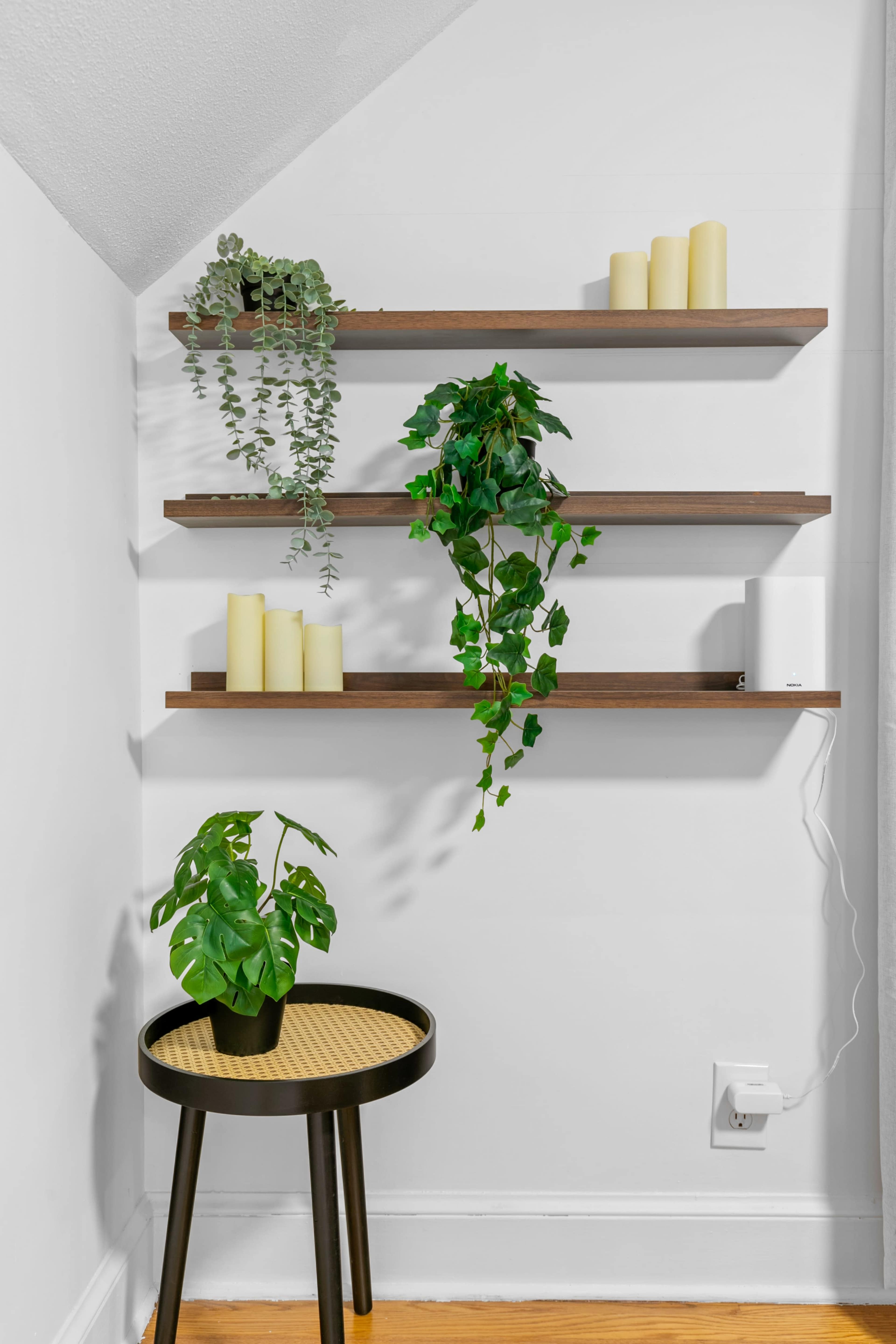 A corner of a room features three wooden shelves adorned with potted plants and candles, alongside a small round table holding a plant.