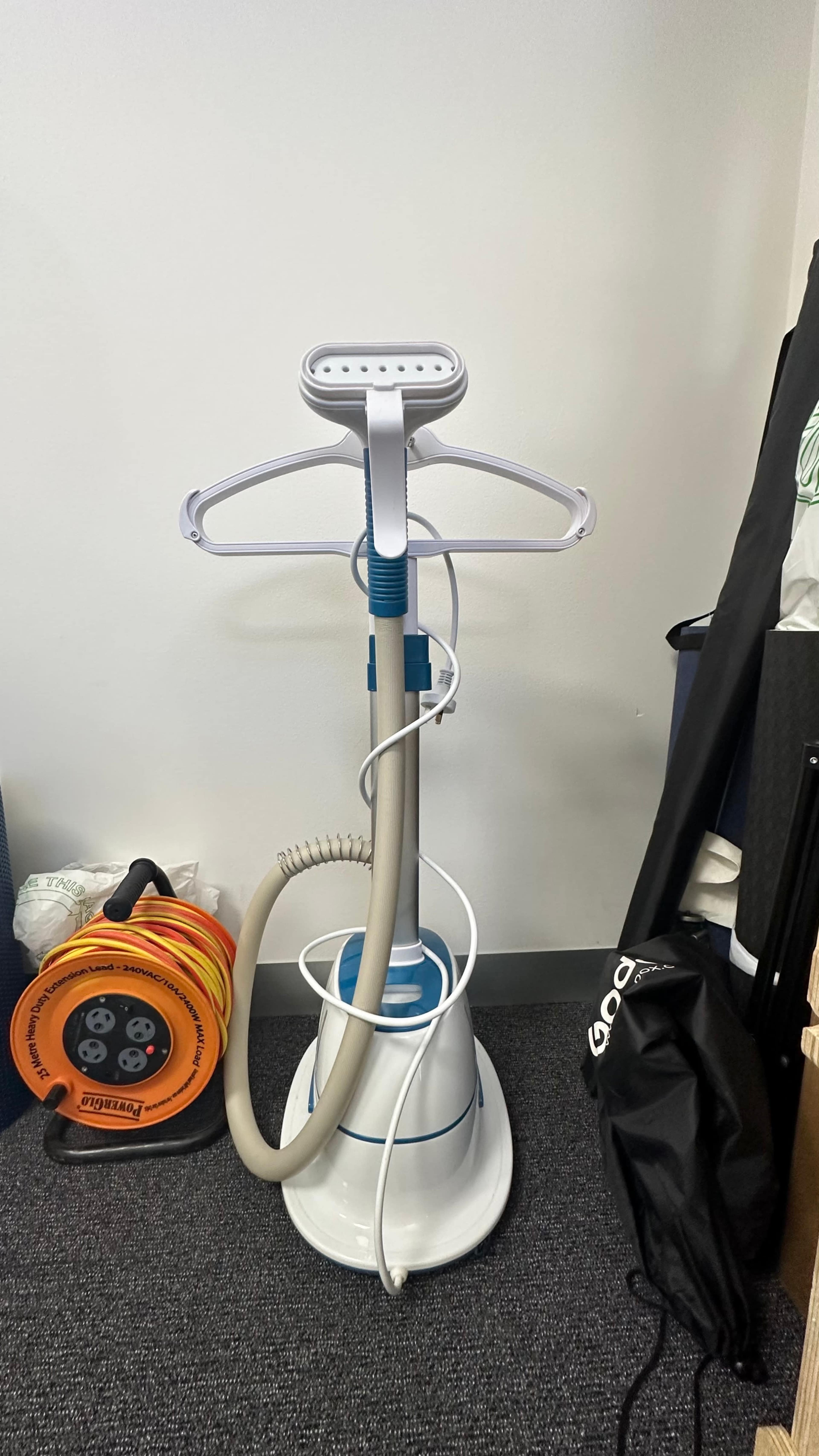 A garment steamer standing on a gray carpet, with a retractable hanger arm and a hose attached, next to a power cord reel and a black bag.