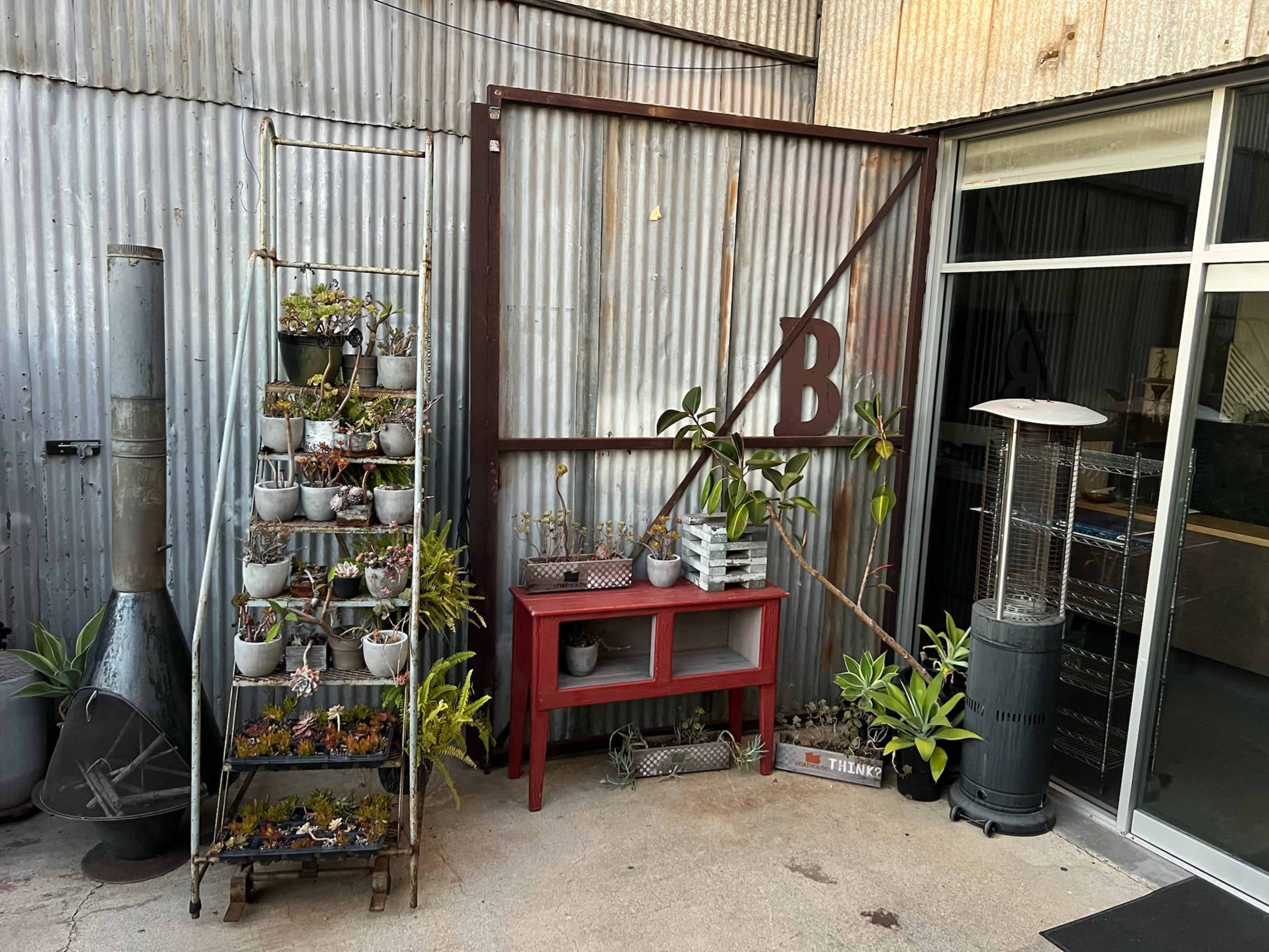 The image shows a small outdoor area with a metal wall, a vertical planter filled with small plants, a red shelving unit, and a patio heater.