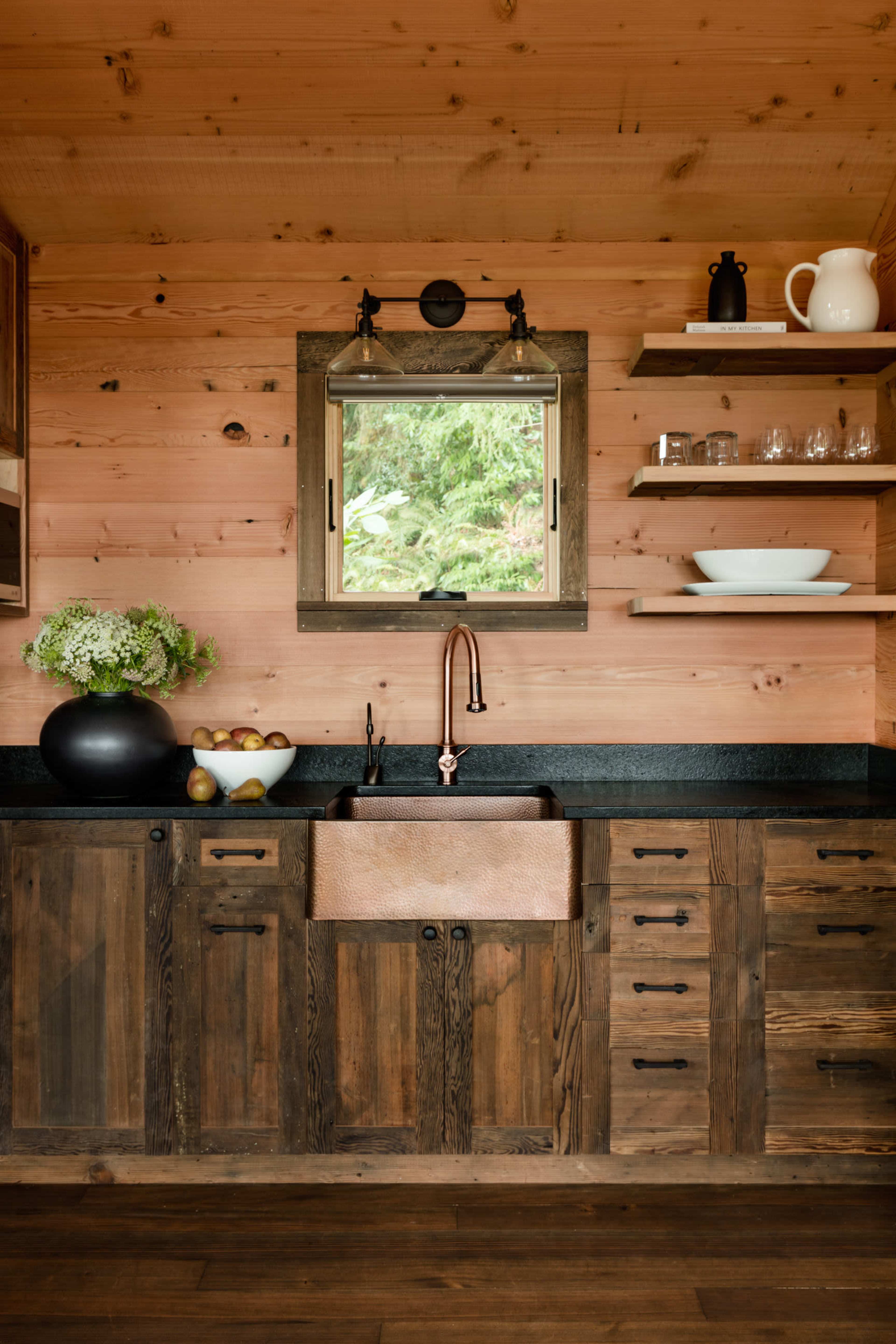 A rustic kitchen with a large copper farmhouse sink beneath a window, wooden cabinetry, and open shelving displaying dishes and decorative items.