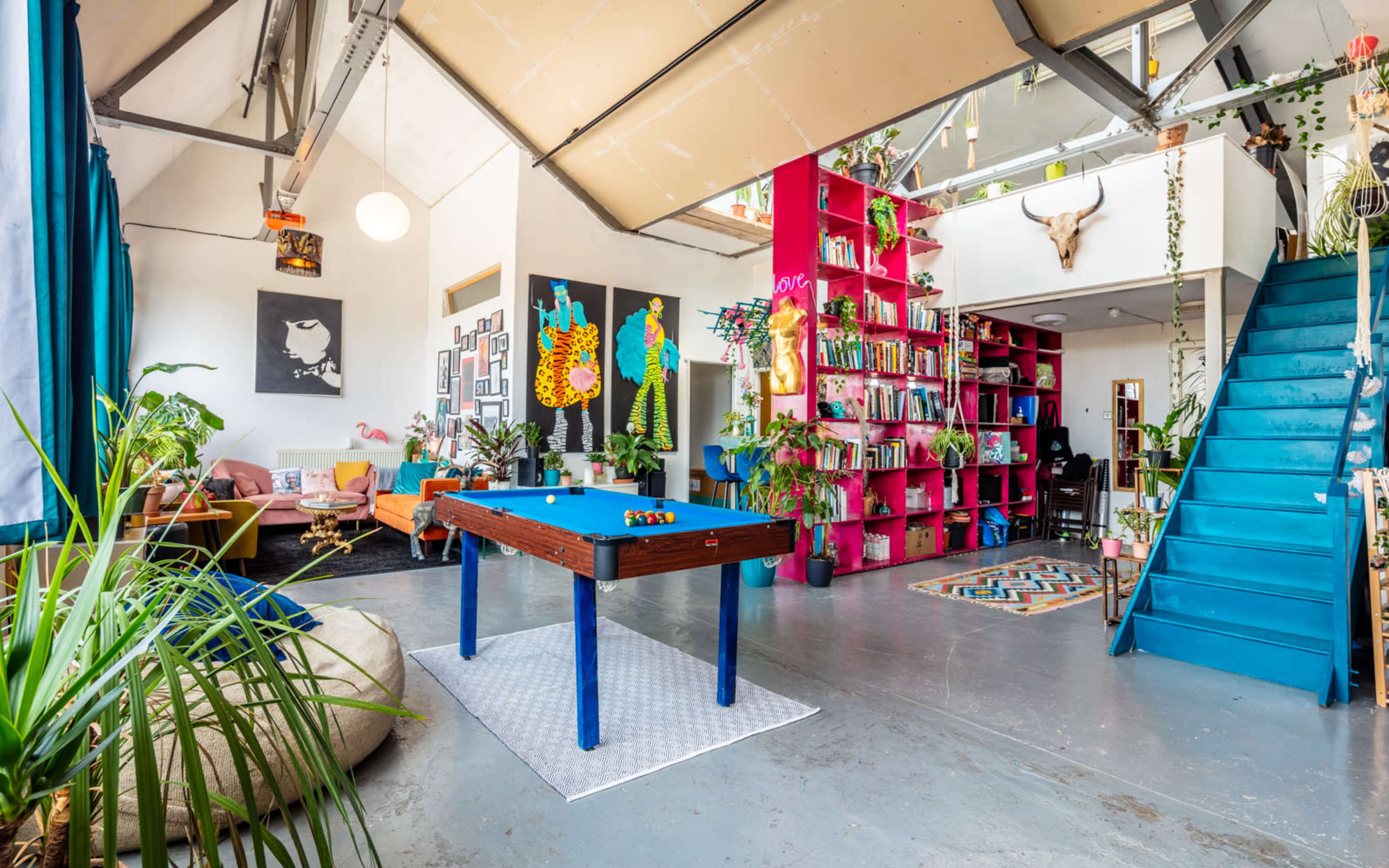 A vibrant interior space with a blue pool table in the center, surrounded by colorful decor, bookshelves, and a staircase leading to an upper level.
