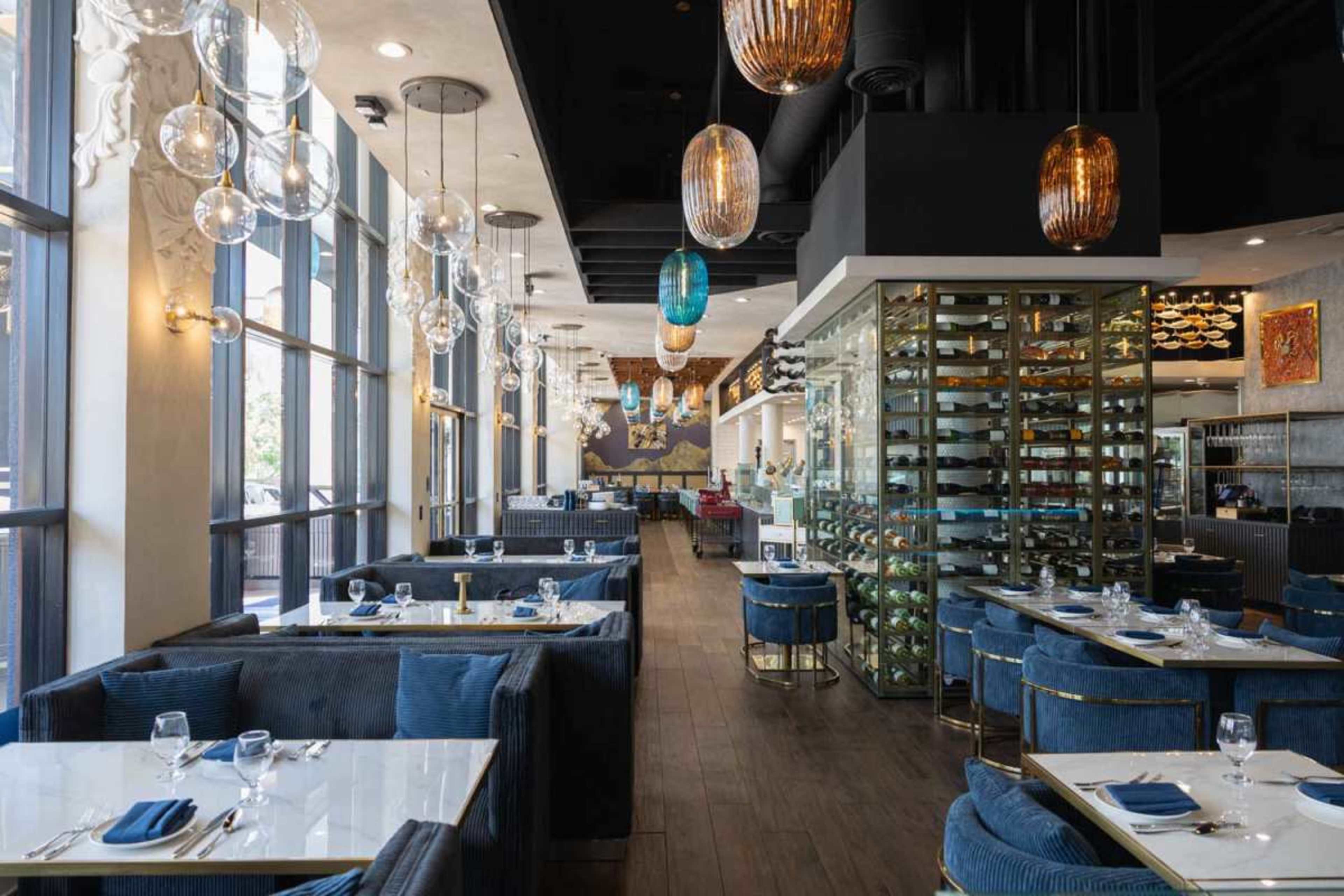 The image shows a modern restaurant interior with blue velvet seating, large windows, and decorative lighting fixtures.