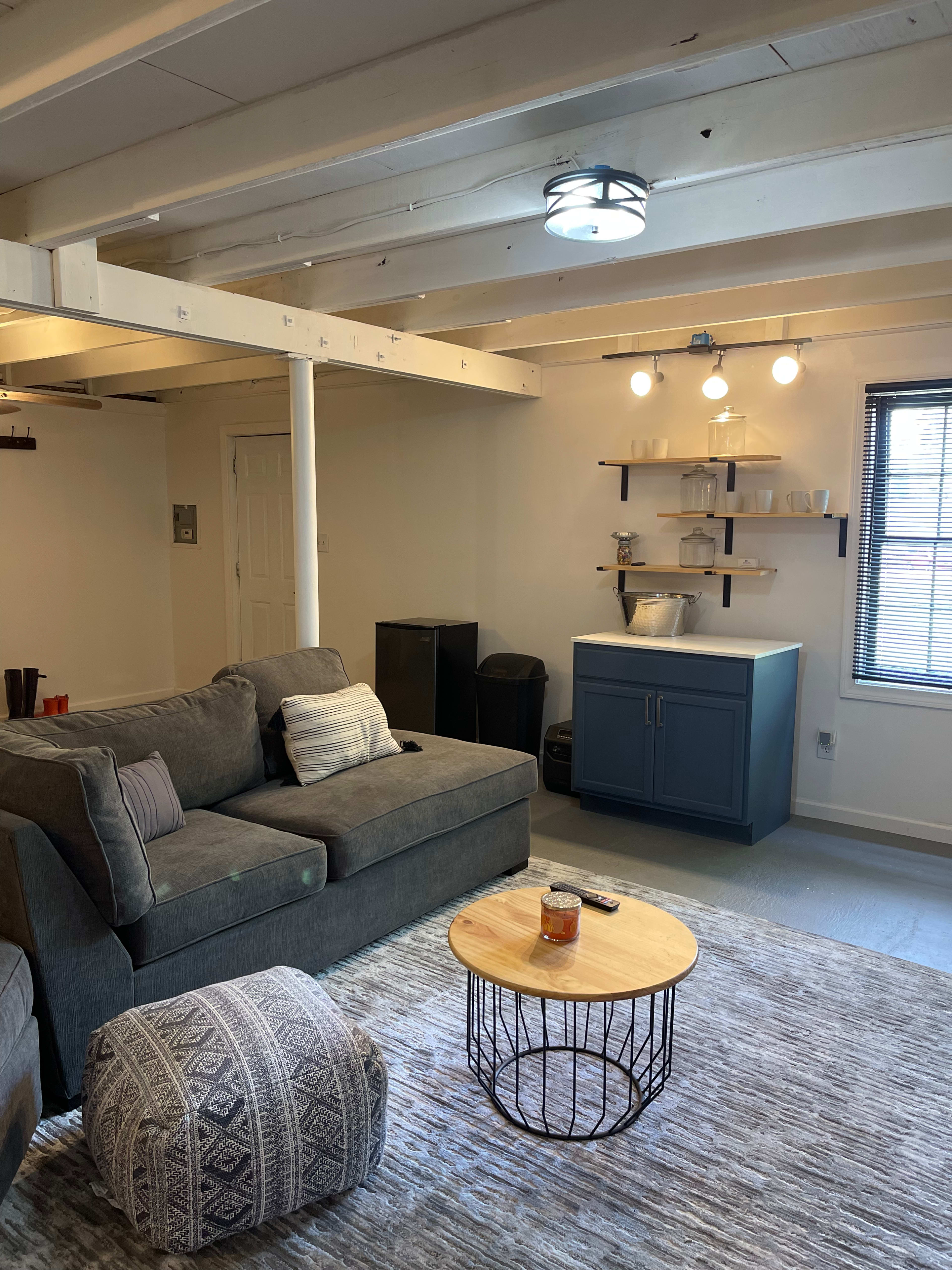 A cozy living area with a gray sectional sofa, a wooden coffee table, a small bar area with a blue cabinet, and a rug underfoot in a well-lit room.