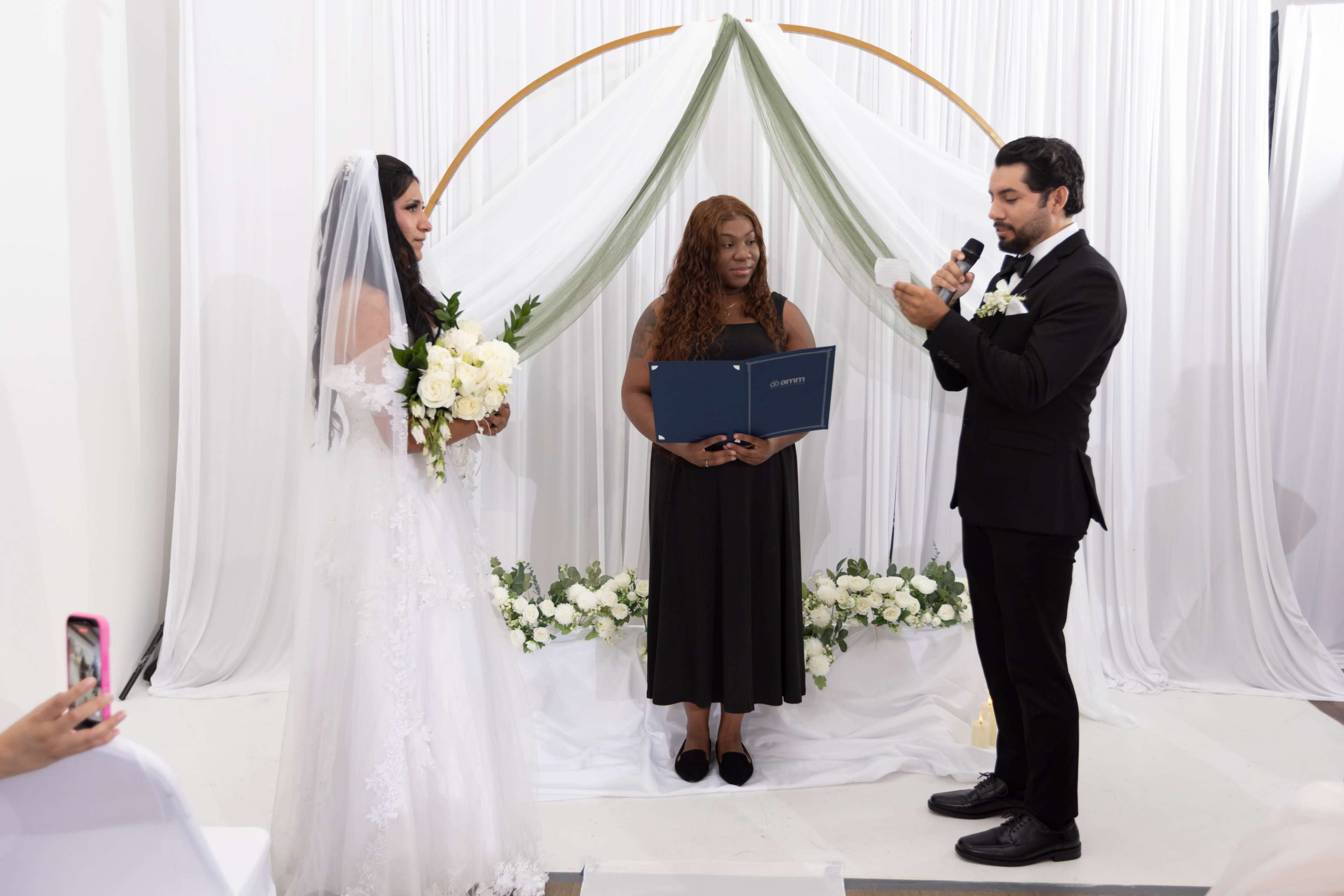 A couple stands under a decorative arch while exchanging vows during their wedding ceremony, with an officiant holding a document in front of them.