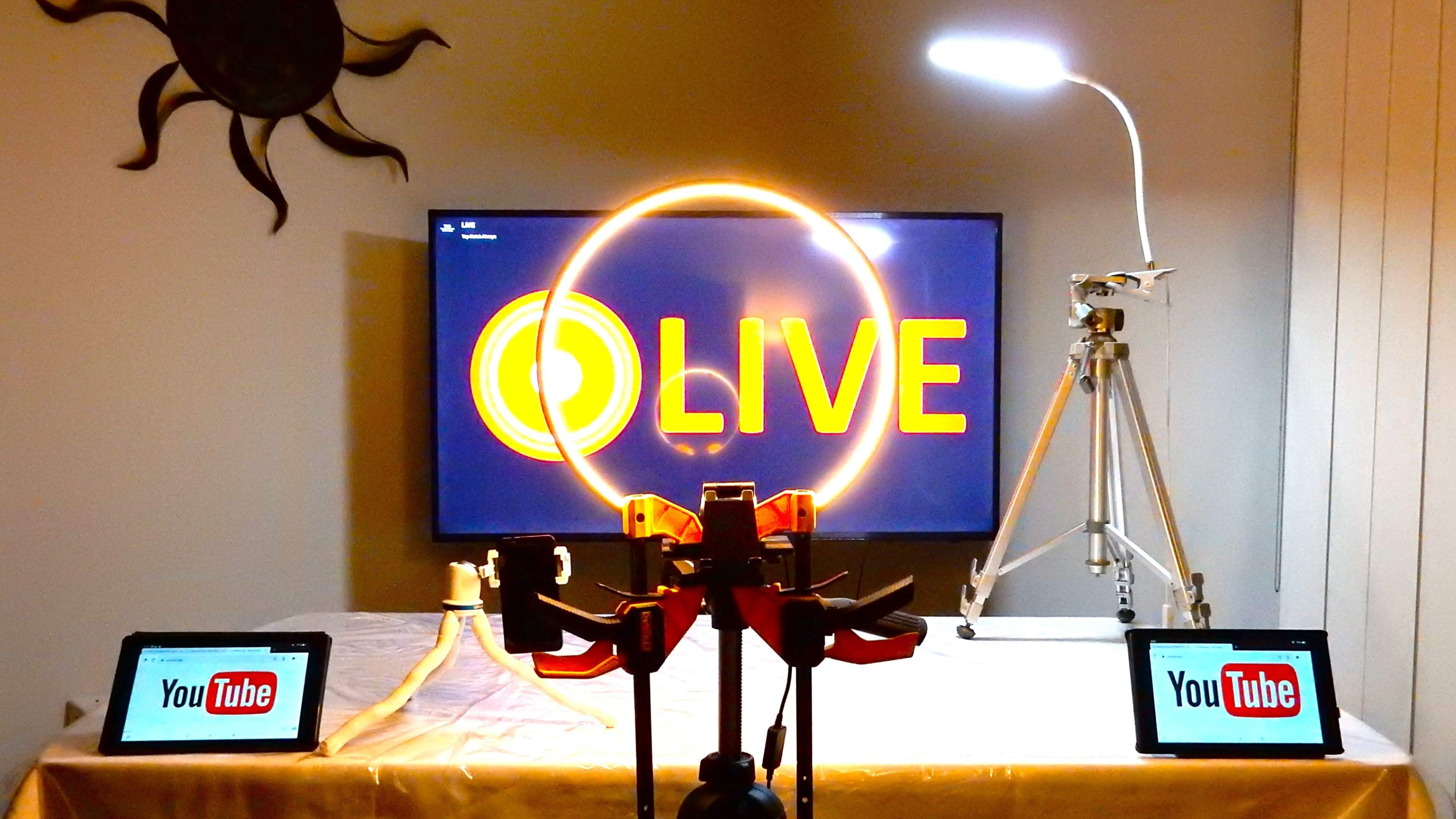 The image shows a setup for a live streaming broadcast, featuring a television displaying "LIVE," a ring light, a tripod, and two tablets with the YouTube logo.