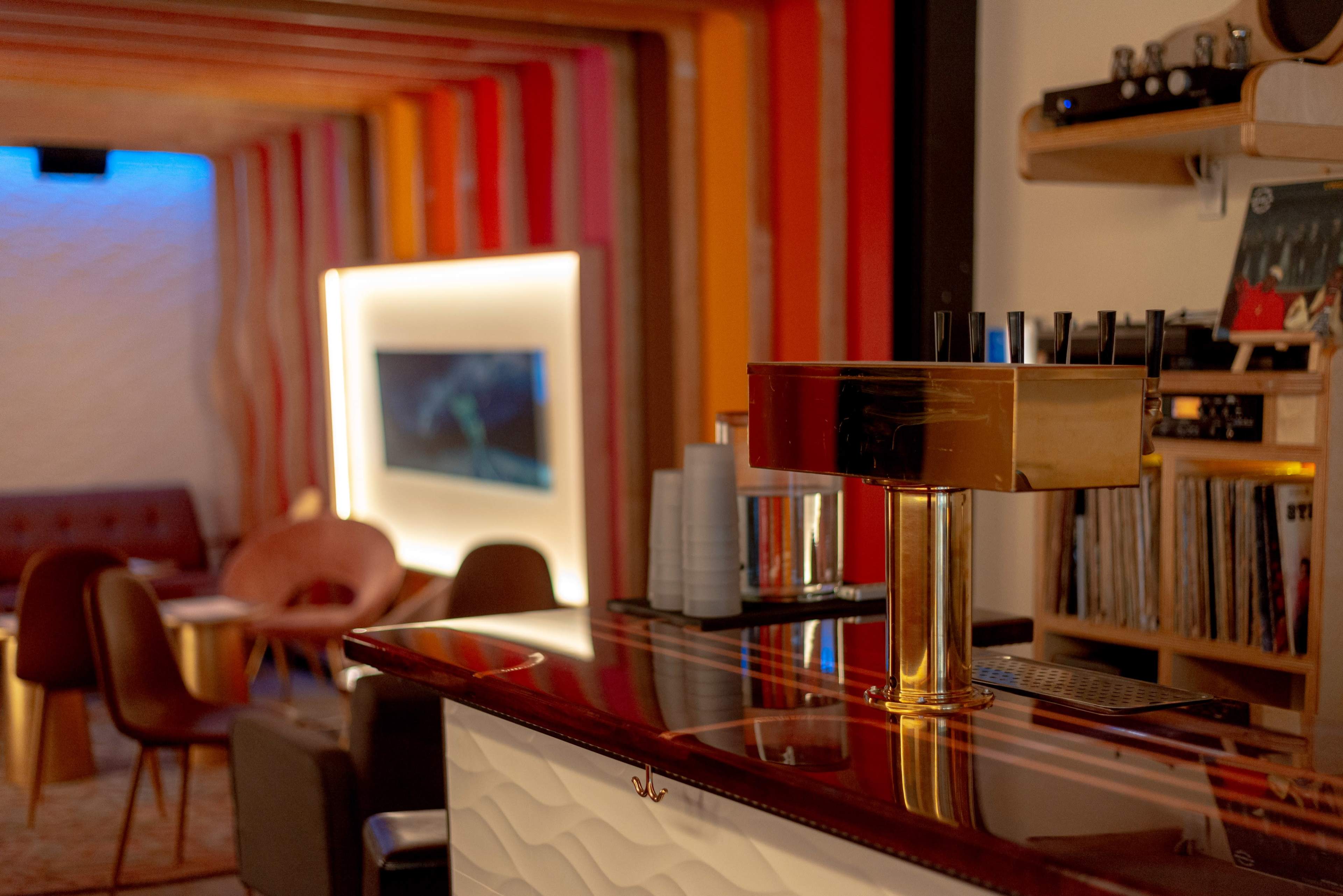 A modern bar with a gold tap stands prominently in the foreground, while colorful walls and comfortable seating create a cozy lounge area in the background.