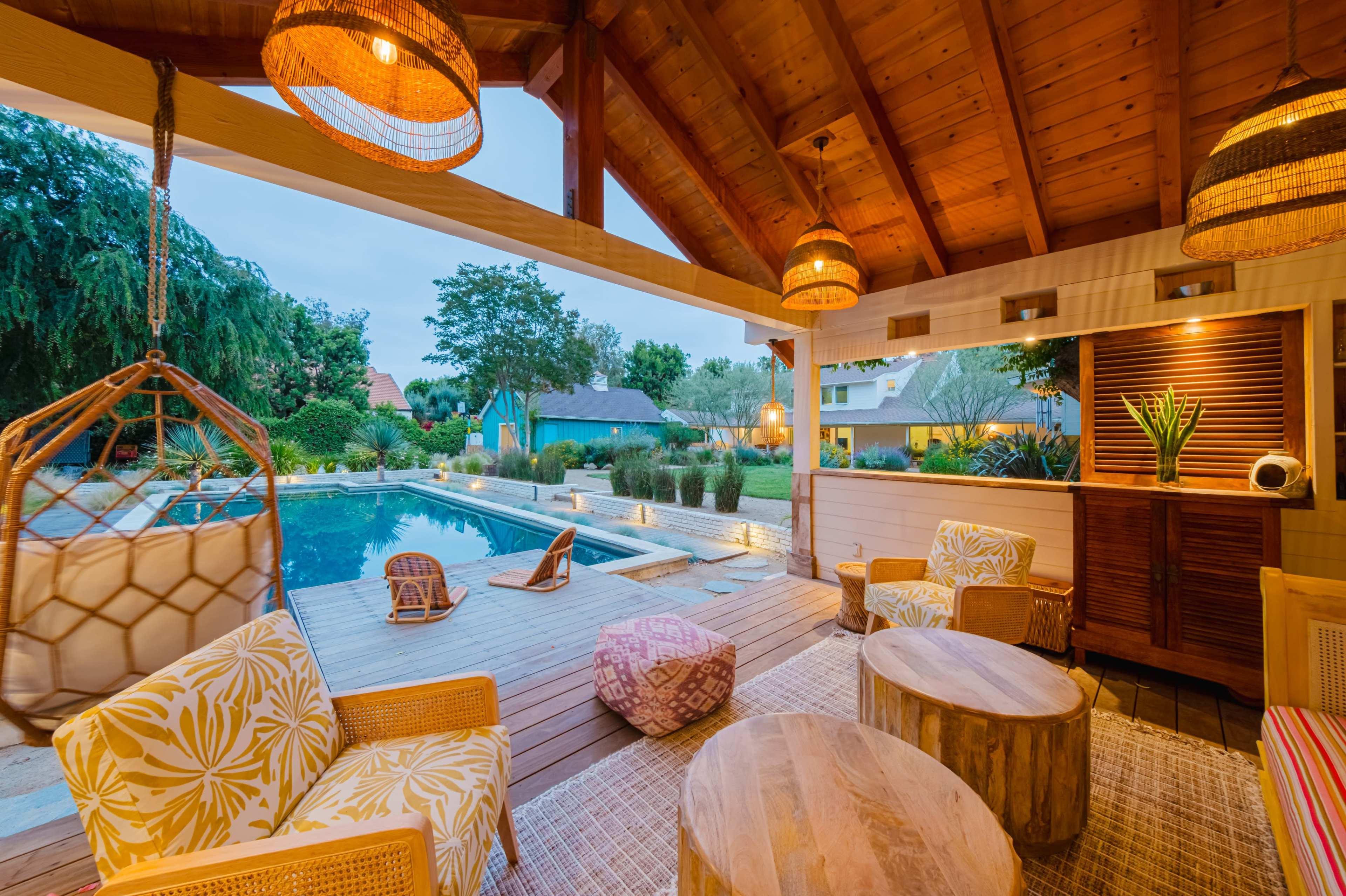 The scene depicts a cozy outdoor space with a pool, furnished with wooden tables and chairs, surrounded by greenery and under a wooden roof with hanging light fixtures.