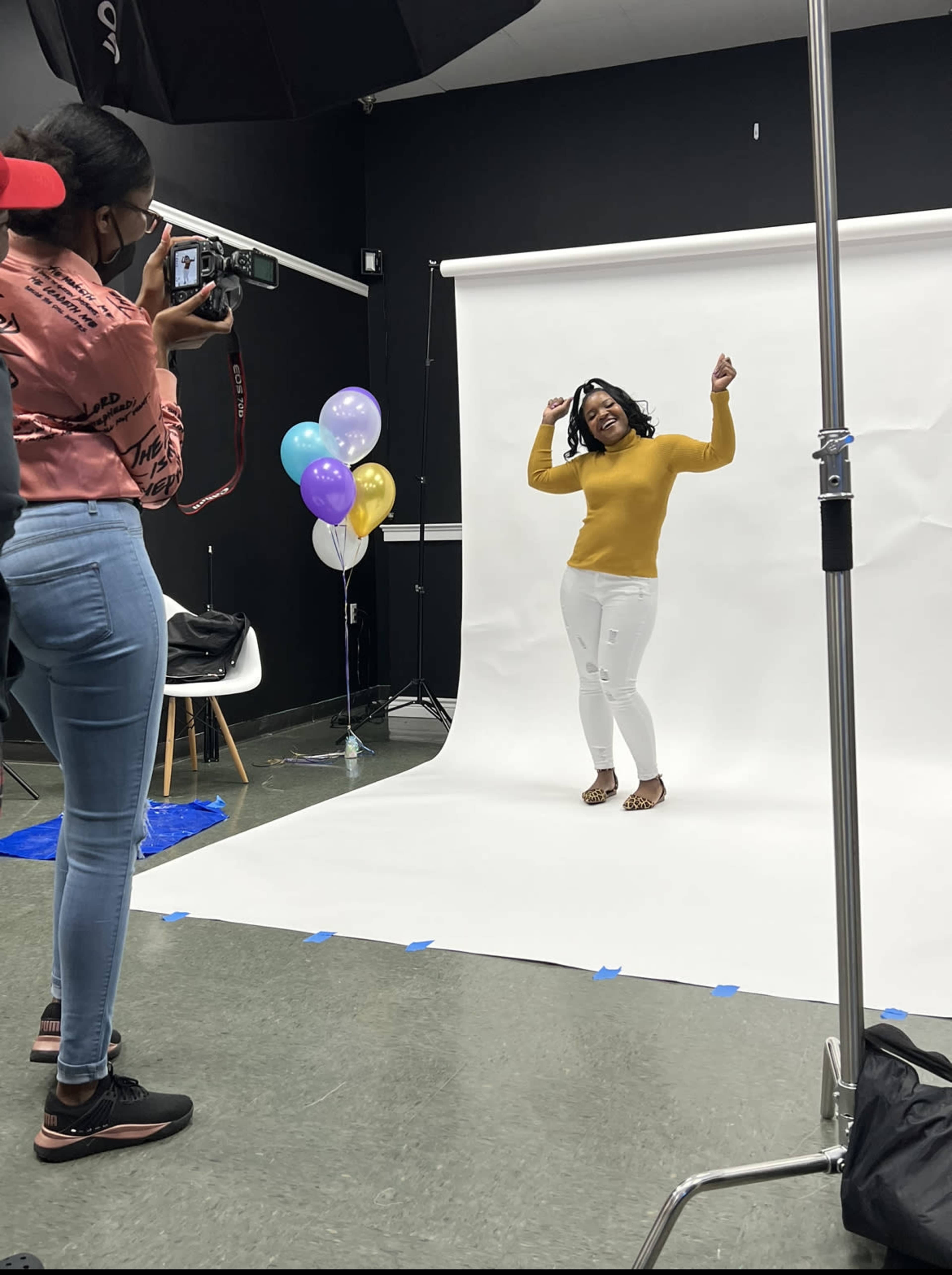 A person wearing a yellow sweater poses playfully in front of a white backdrop while another individual captures the moment with a camera.