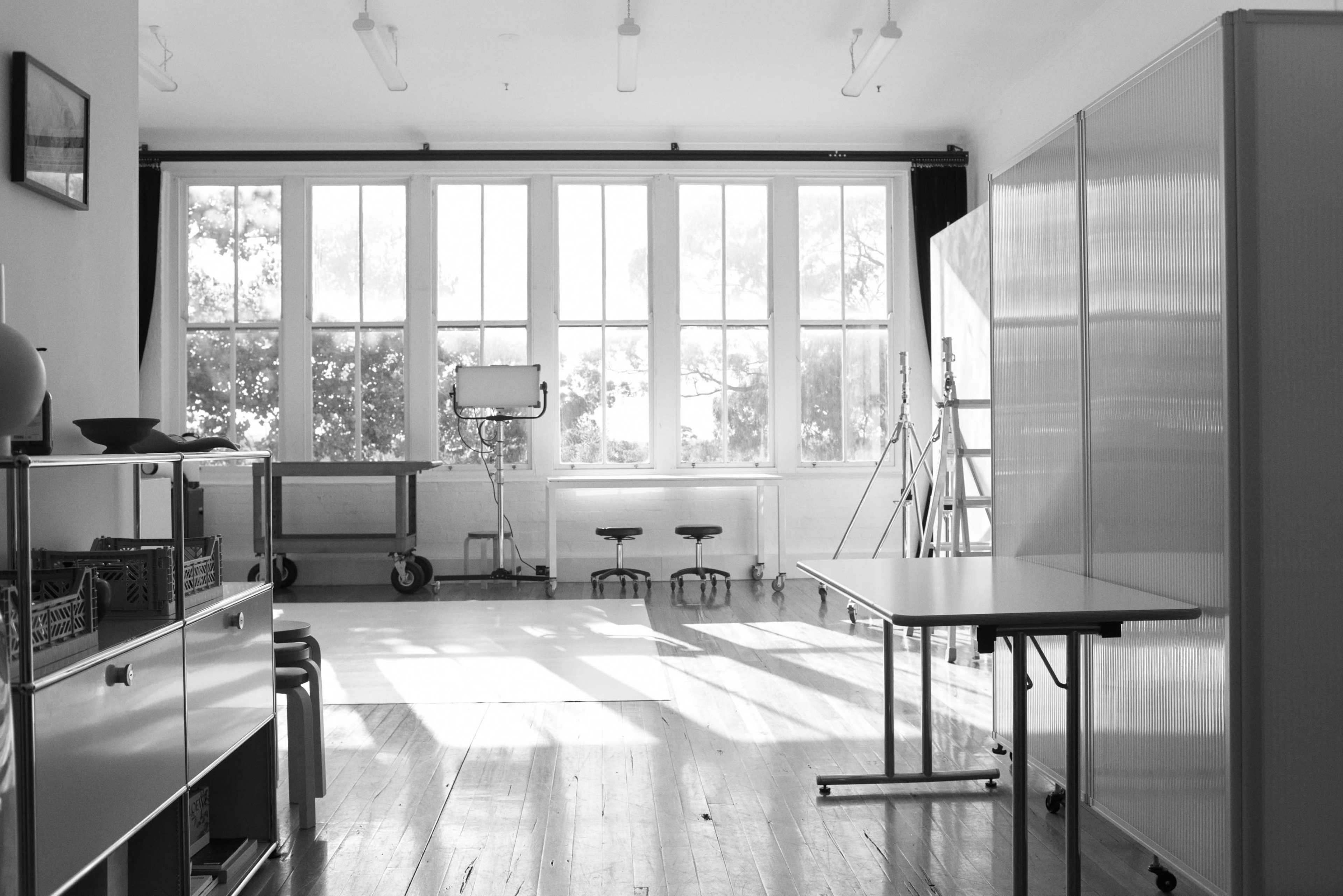 A spacious, empty room with large windows, light wood flooring, and a few pieces of furniture including tables and stools.