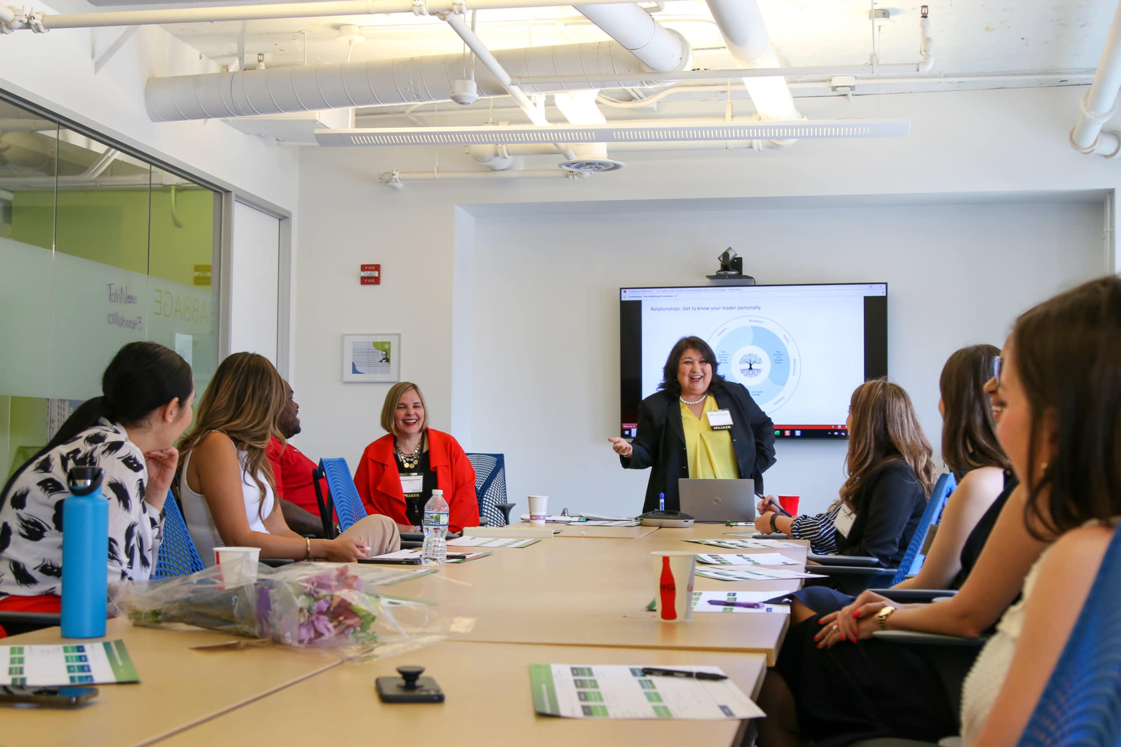 A group of women is seated around a conference table, actively participating in a presentation being given by a speaker, with a screen displaying information in the background.