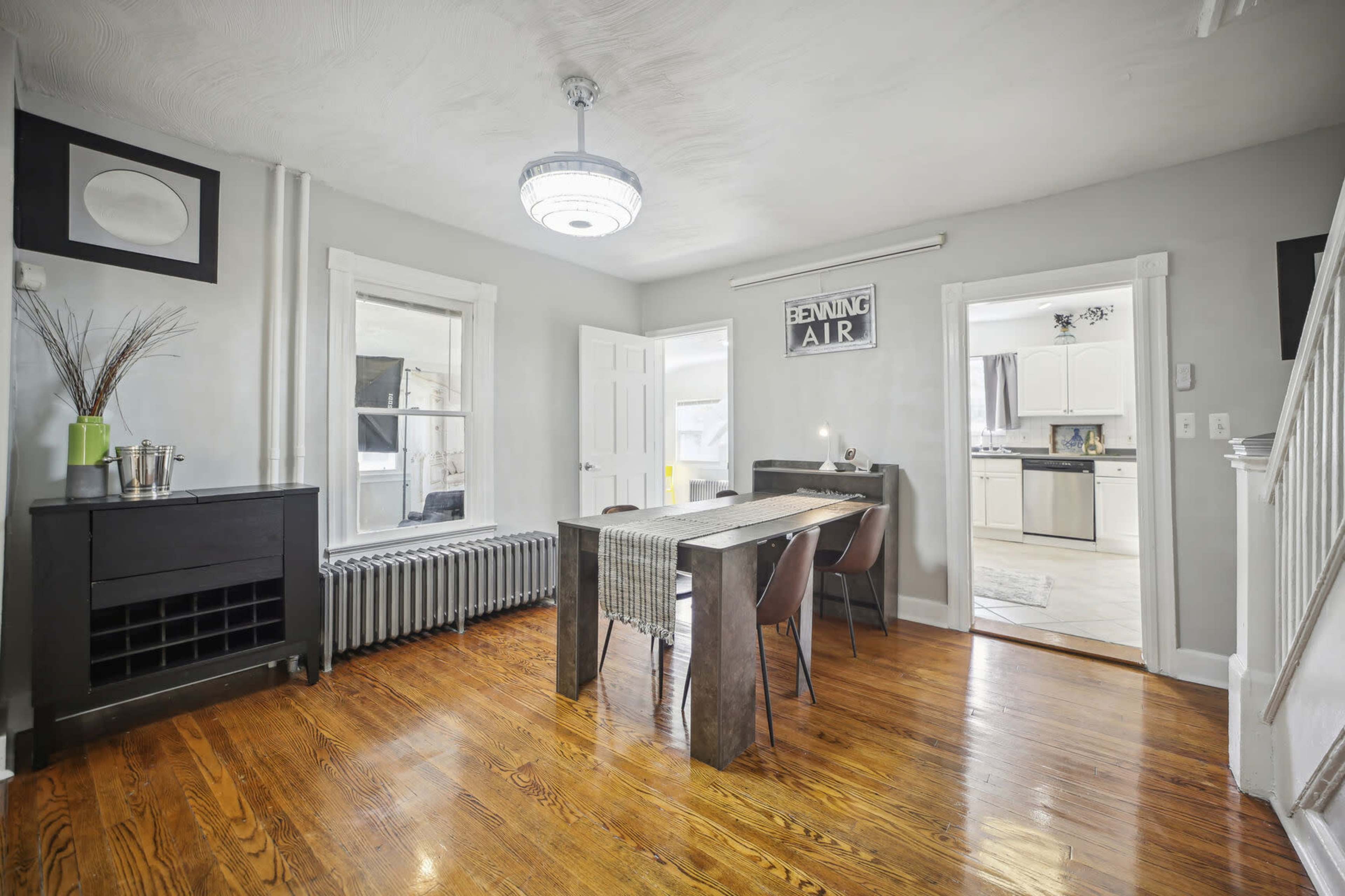 A dining area with a wooden table, four chairs, and a radiator, adjacent to a kitchen and two windows.