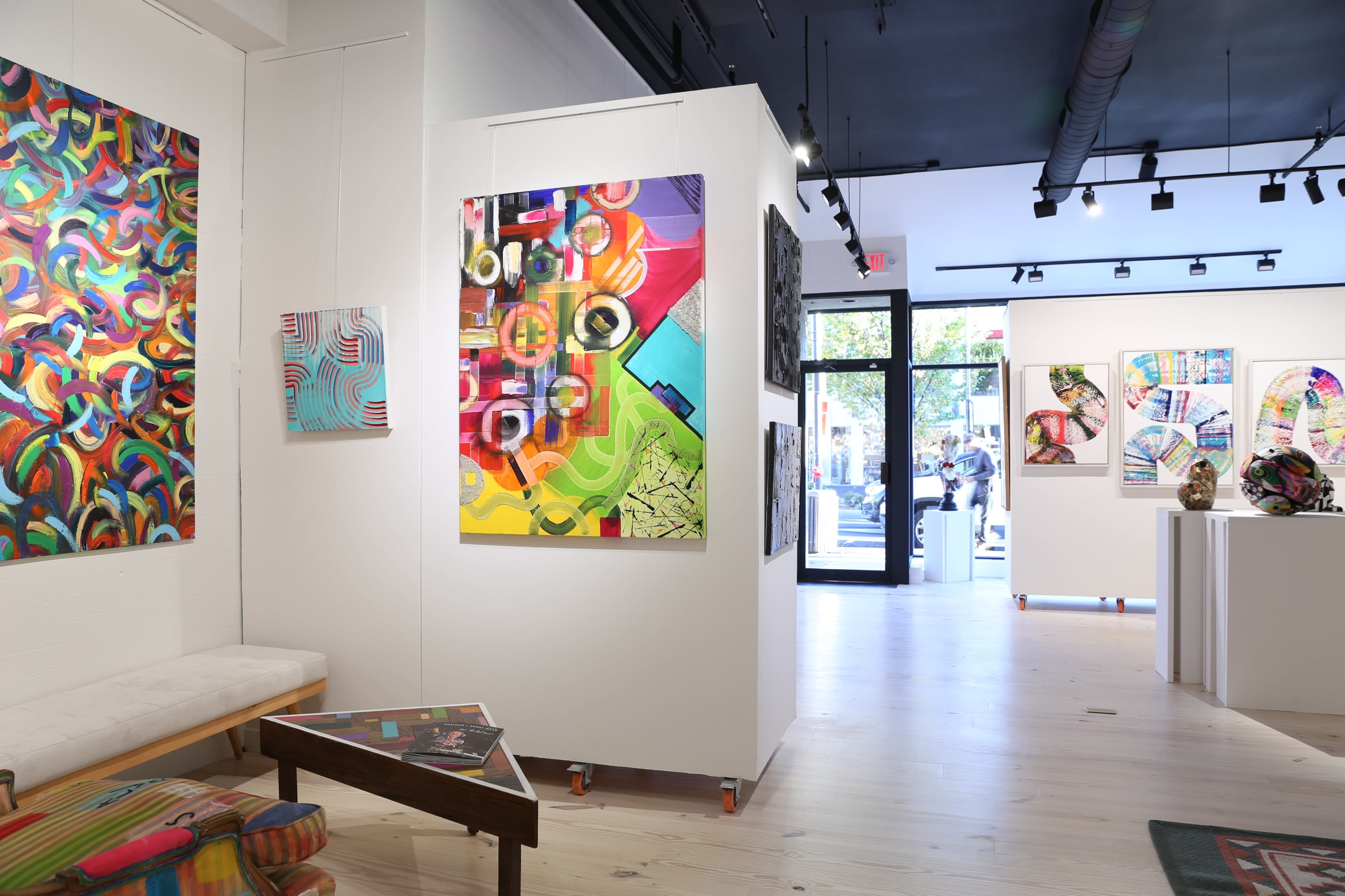 A contemporary art gallery featuring colorful abstract paintings displayed on the white walls, with a bright entrance leading to the outside.