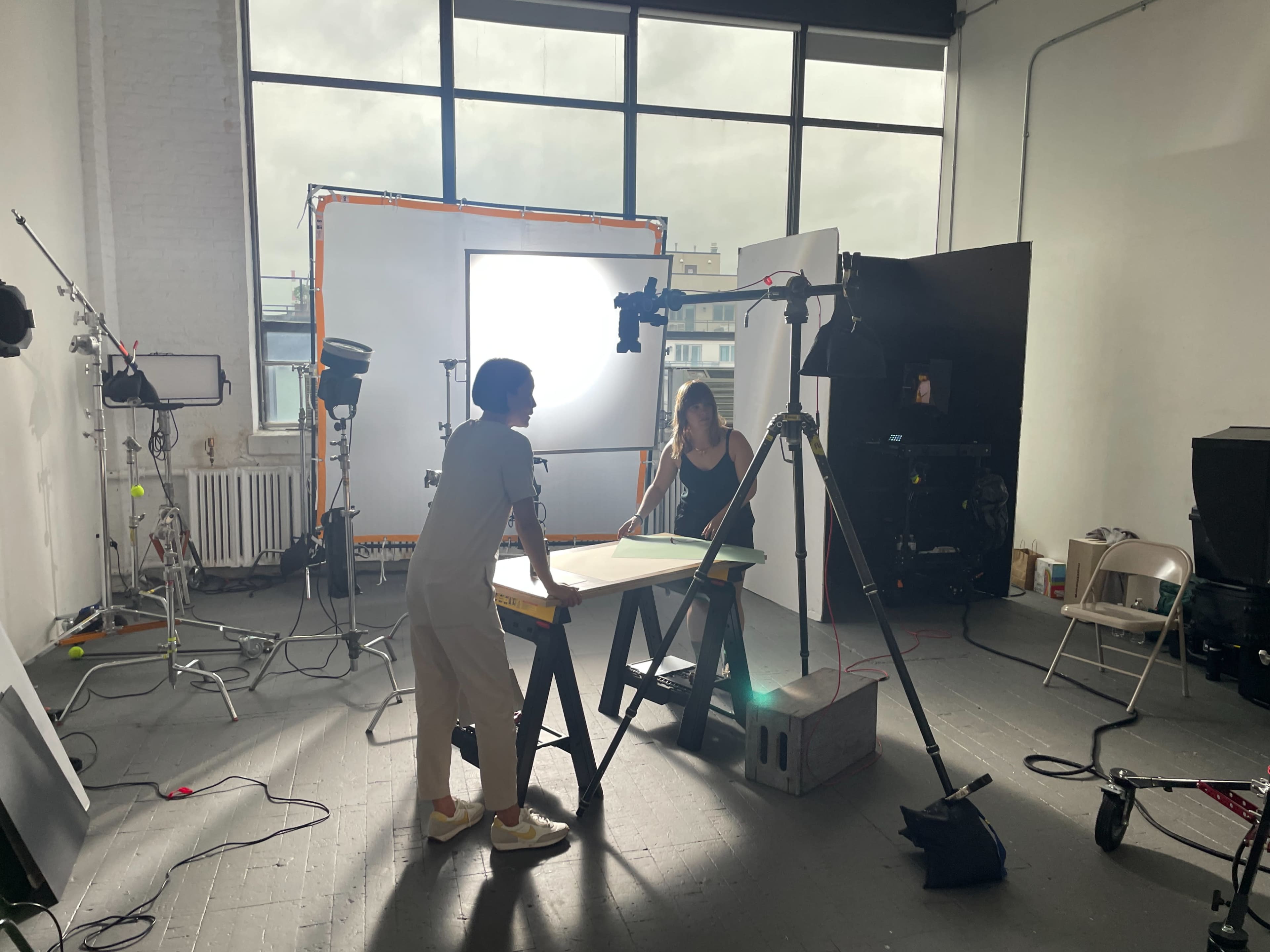 Two individuals are working at a table in a photography studio filled with lights and equipment.