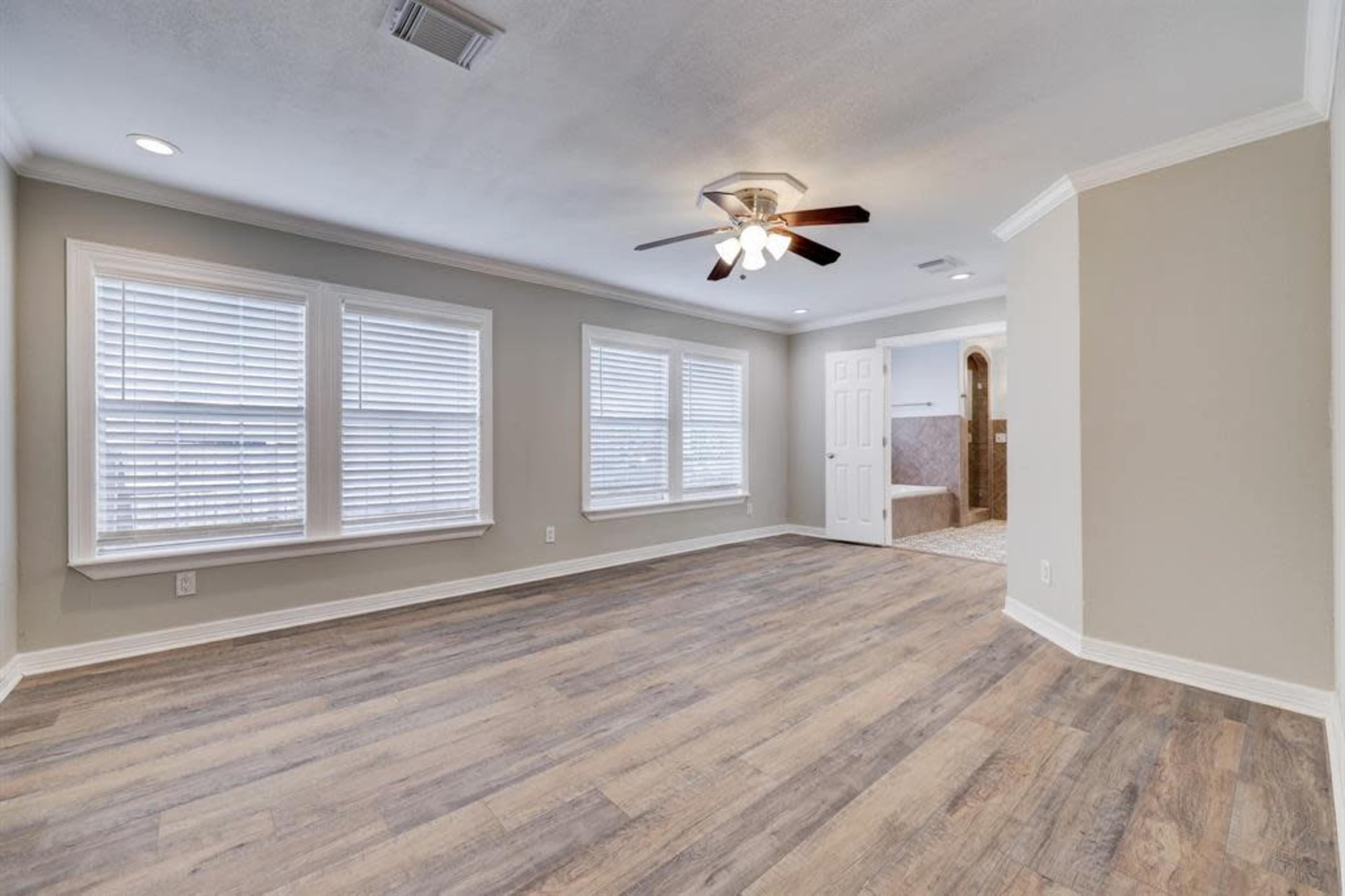 The image shows a spacious, light-filled living room with wooden flooring, multiple windows, and a ceiling fan, adjacent to a bathroom area.