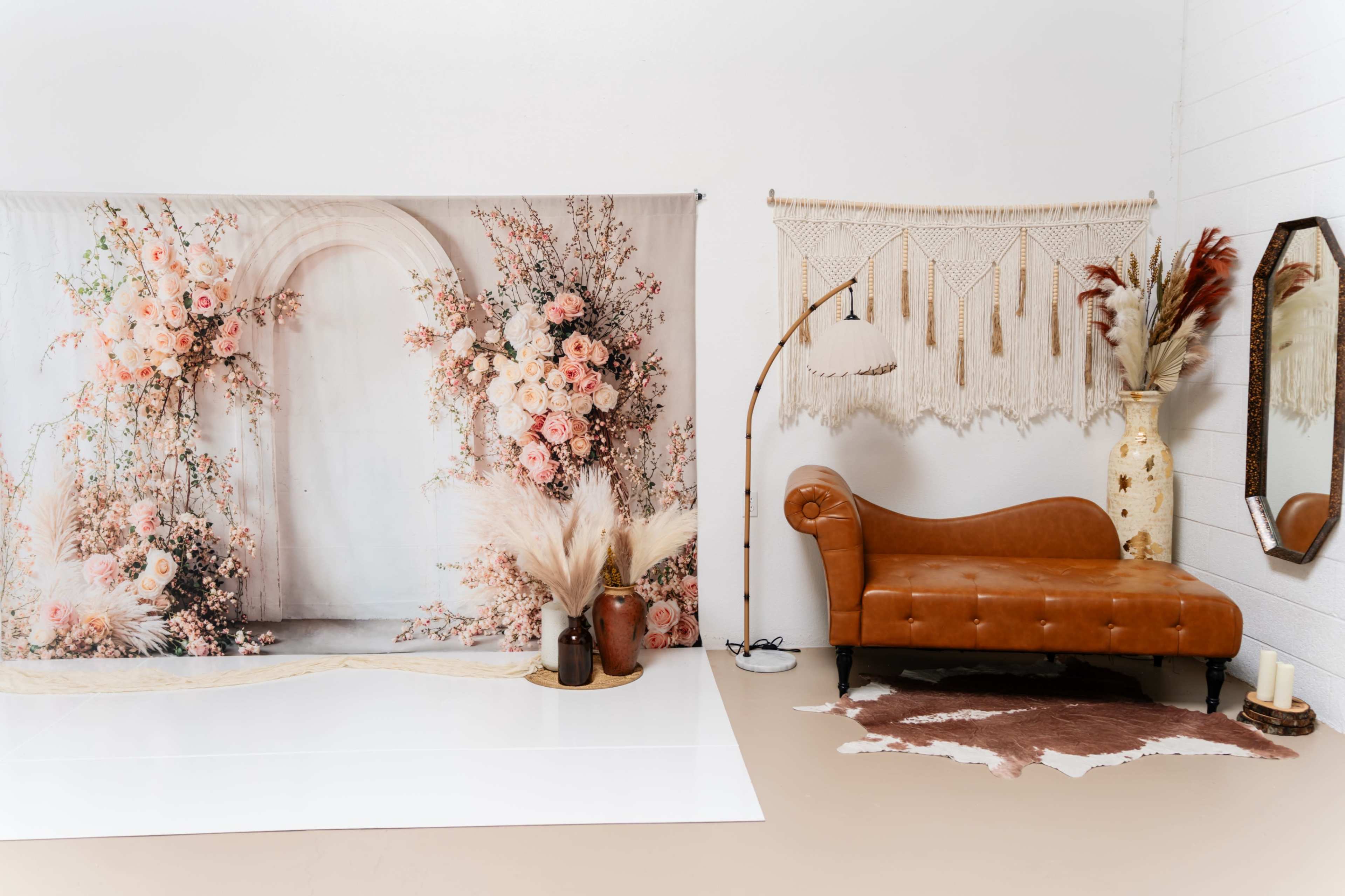 The image features a decorated studio space with a floral backdrop, a tan leather couch, a macramé wall hanging, and decorative vases.