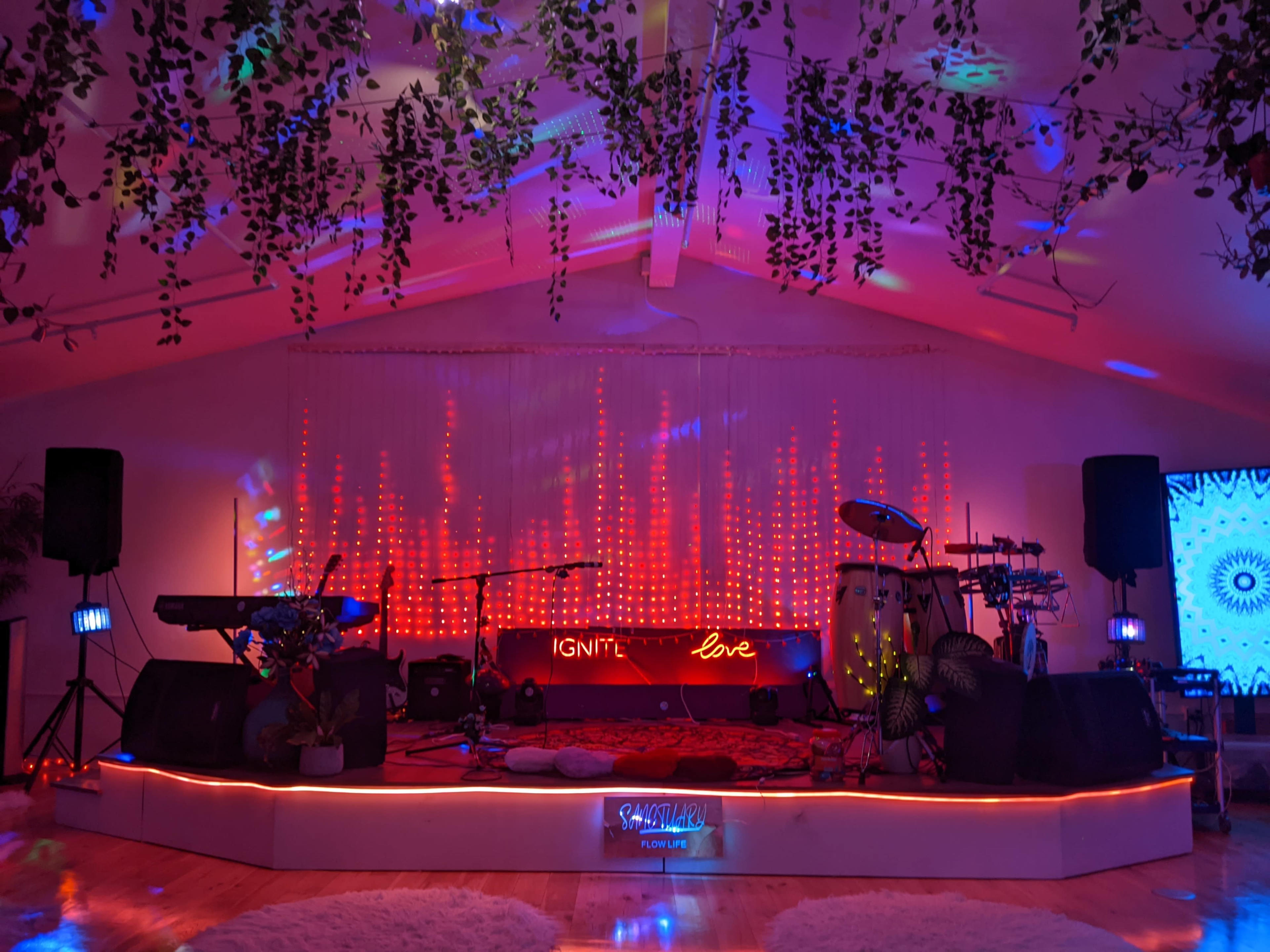 The image shows a stage setup with musical instruments, colorful lighting, and decorative plants overhead in a venue.