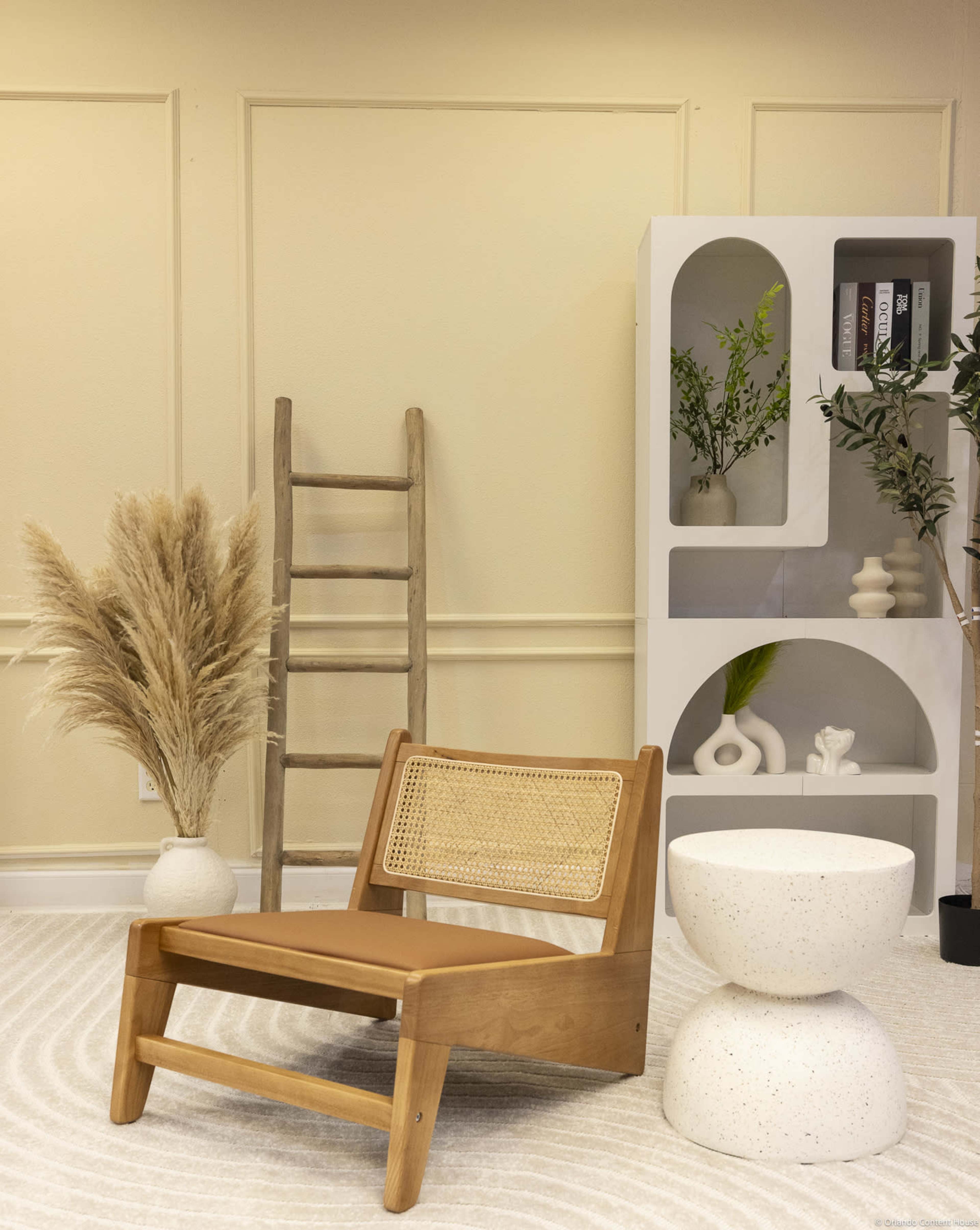 The scene features a minimalist room with a wooden chair, a circular stone table, a decorative ladder, and potted plants arranged against a light-colored wall.