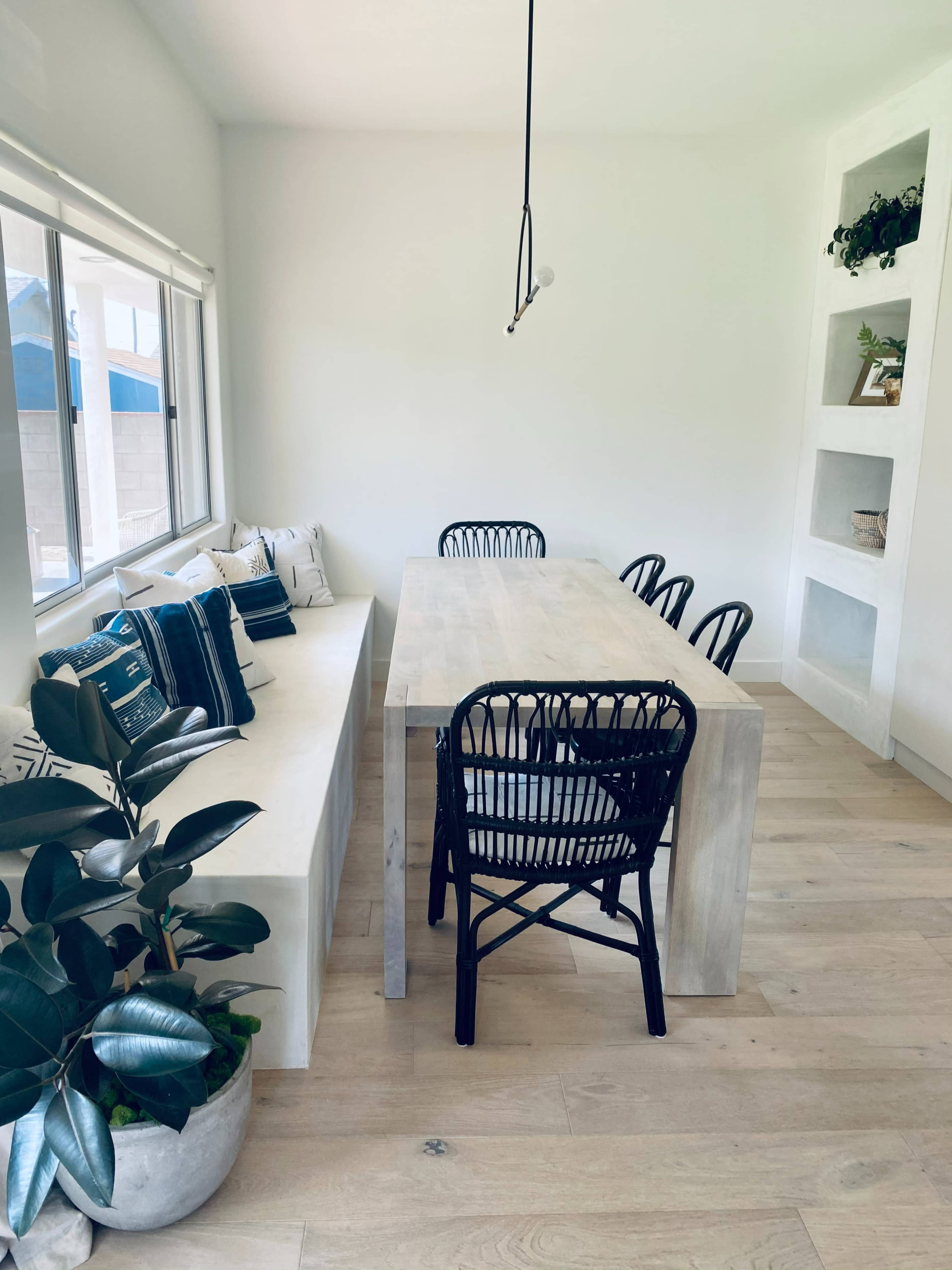 The image shows a modern dining area with a minimalist wooden table surrounded by black chairs, a built-in shelf, and a bench seat lined with decorative pillows.