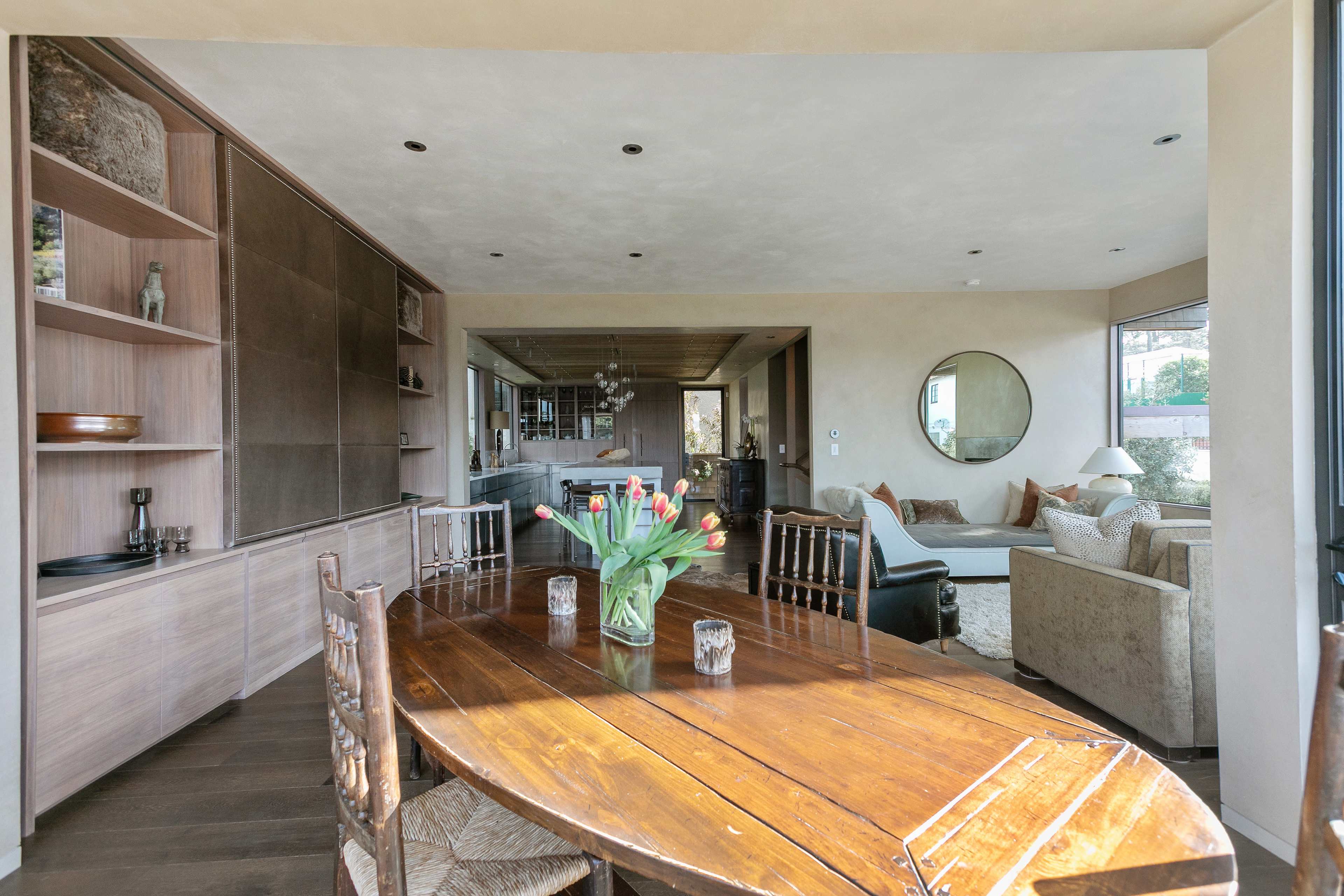 A wooden dining table with a vase of tulips is set in a modern, open-concept living space that includes a kitchen and a sitting area.