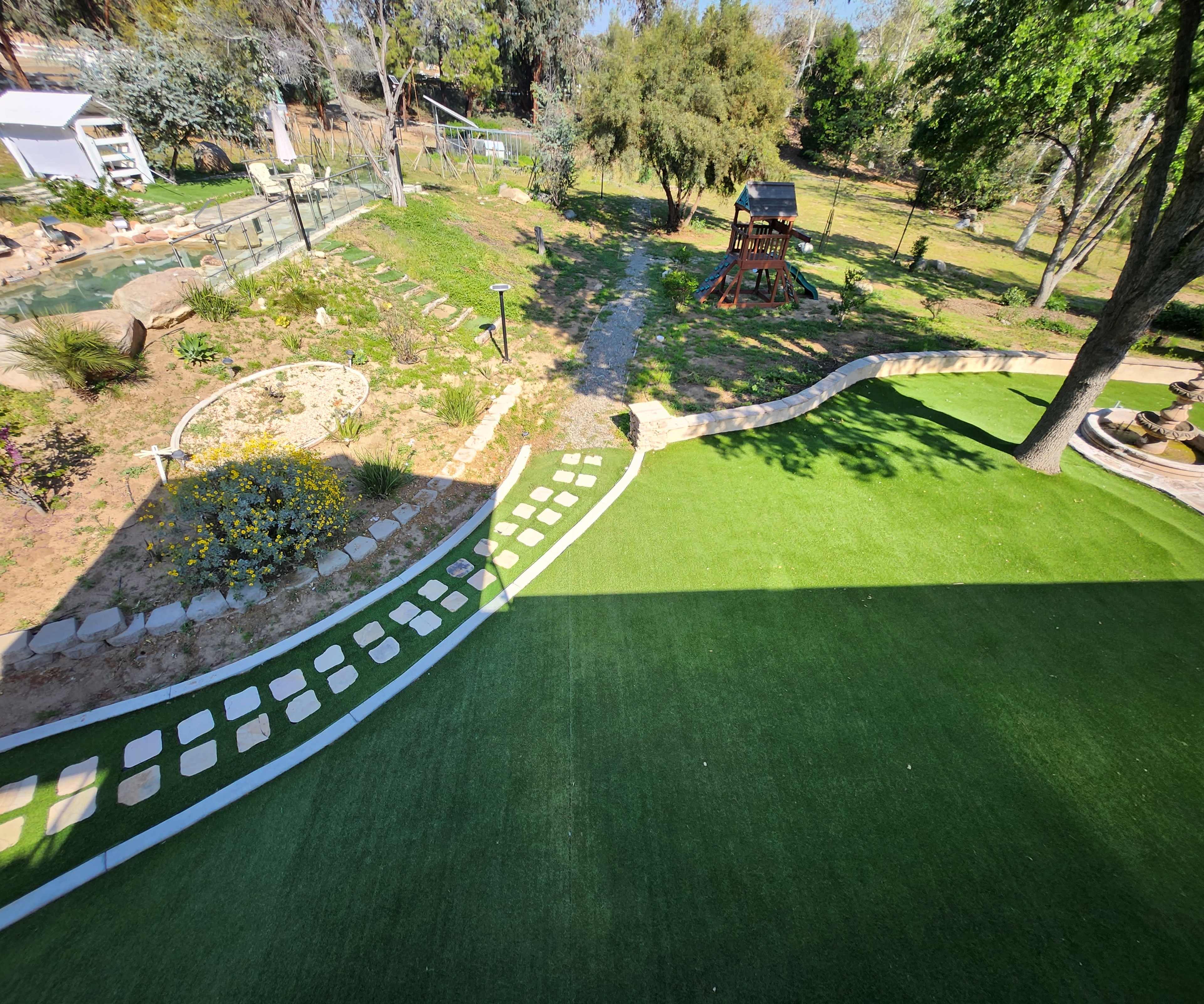 A landscaped backyard featuring artificial turf, a winding path of stepping stones, a play structure, and various plants and trees.