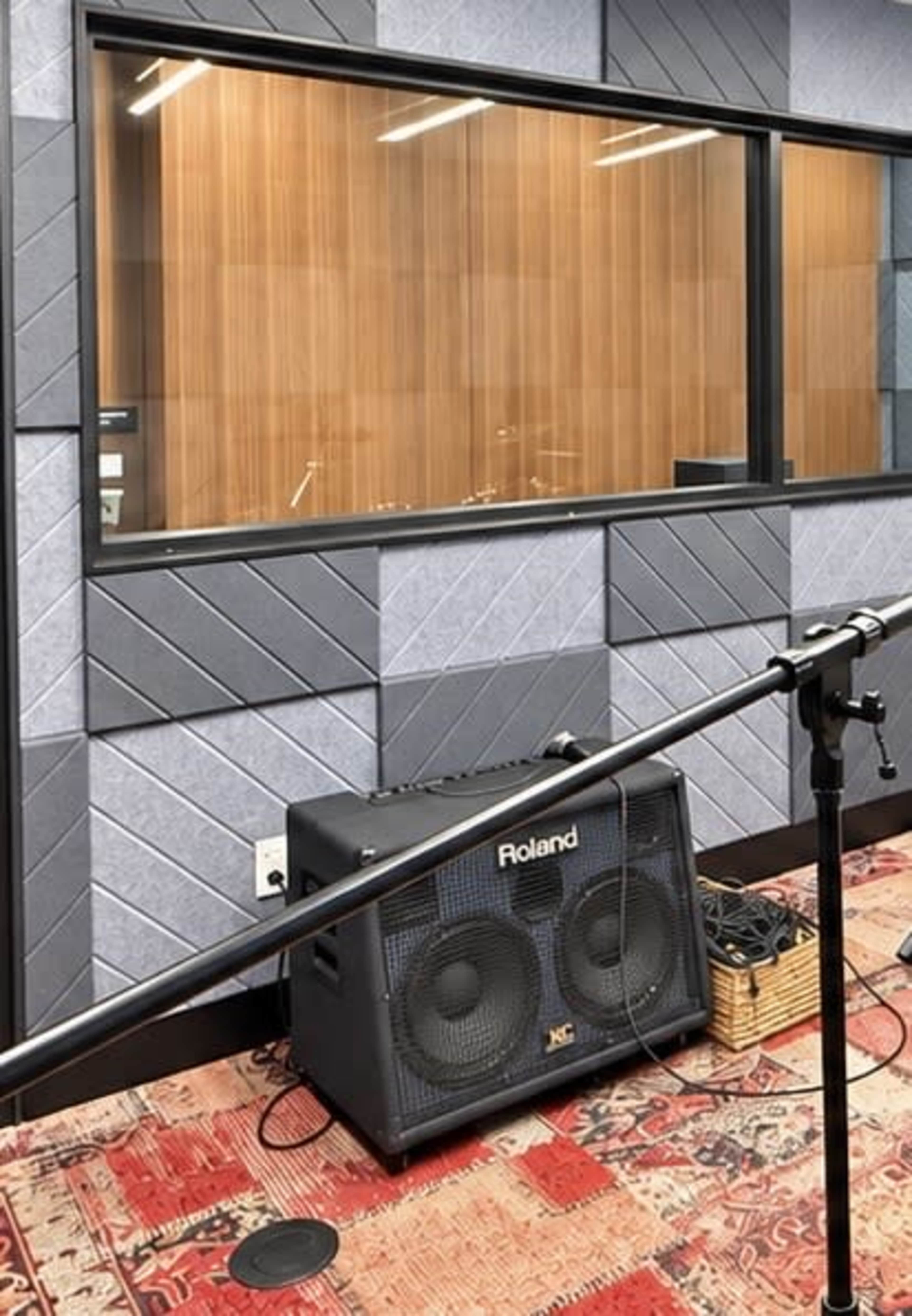 The image shows a sound studio with a glass window, a large amplifier on the floor, and a microphone stand positioned nearby.