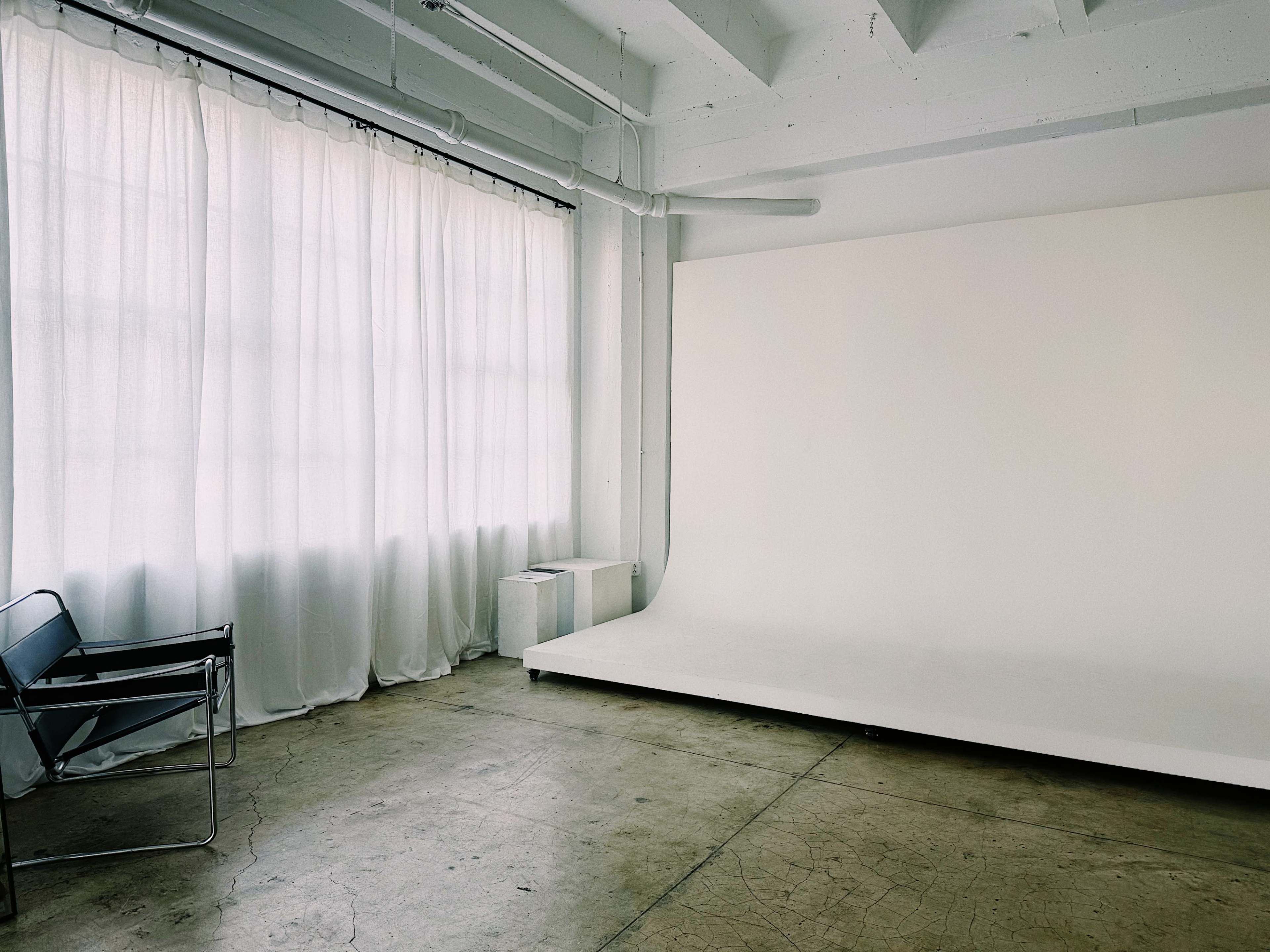 The image shows a spacious room with large windows covered by sheer curtains, featuring an empty white backdrop and a minimalist chair.