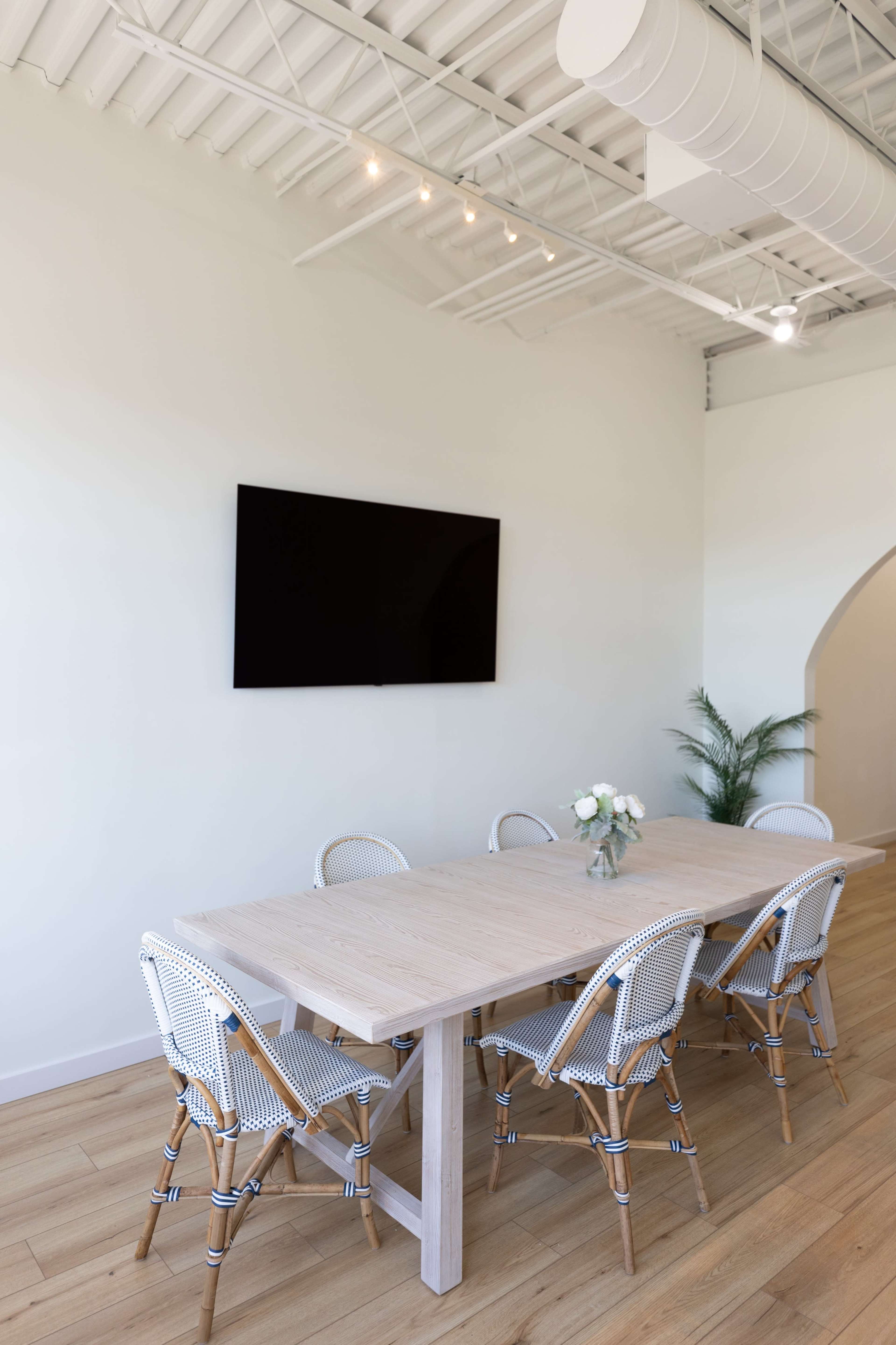 A spacious meeting room features a wooden table surrounded by six chairs and a wall-mounted television.