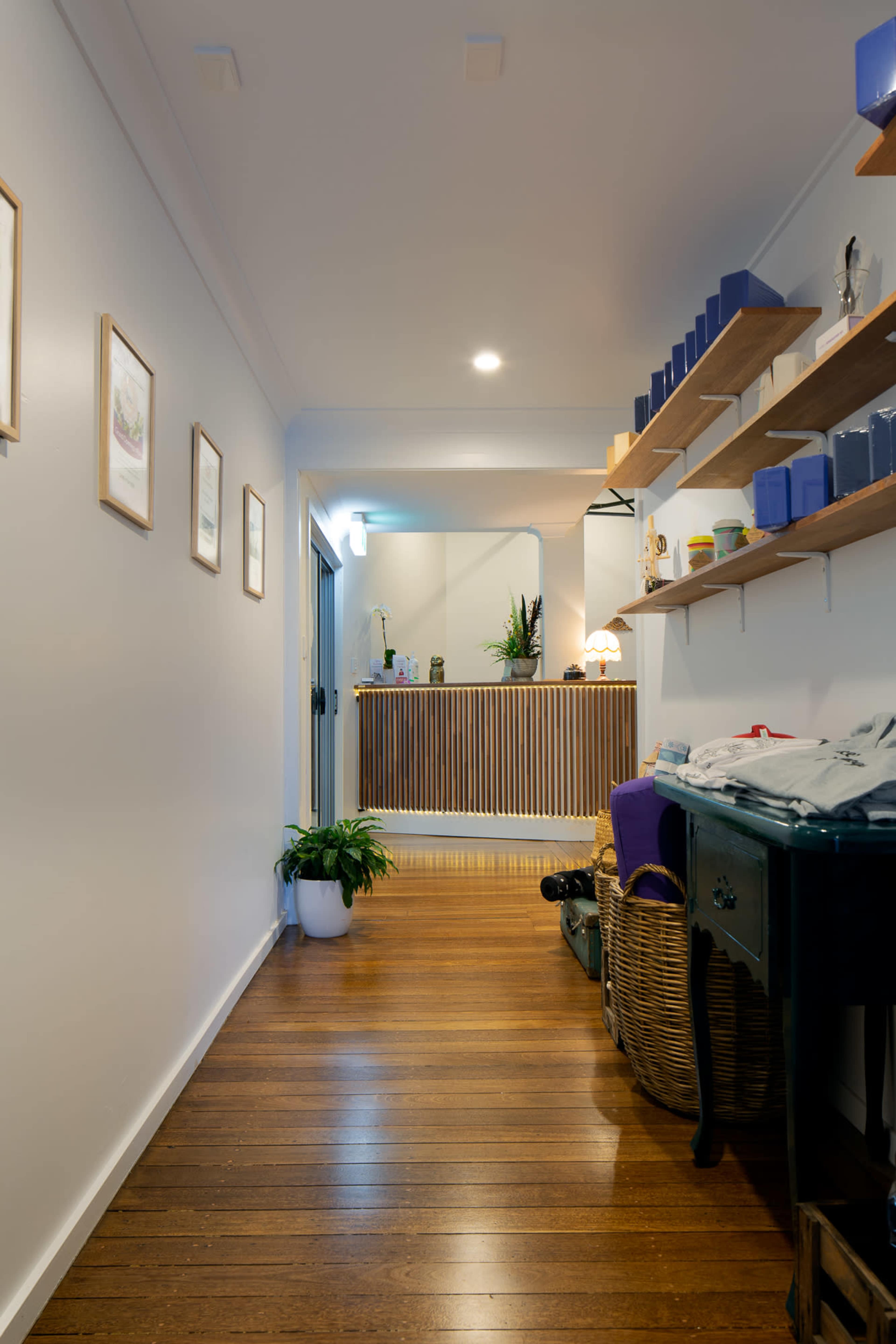 A hallway with wooden flooring, lined with framed pictures on one side and shelves holding various items on the other, leading to a reception area with a wooden counter.