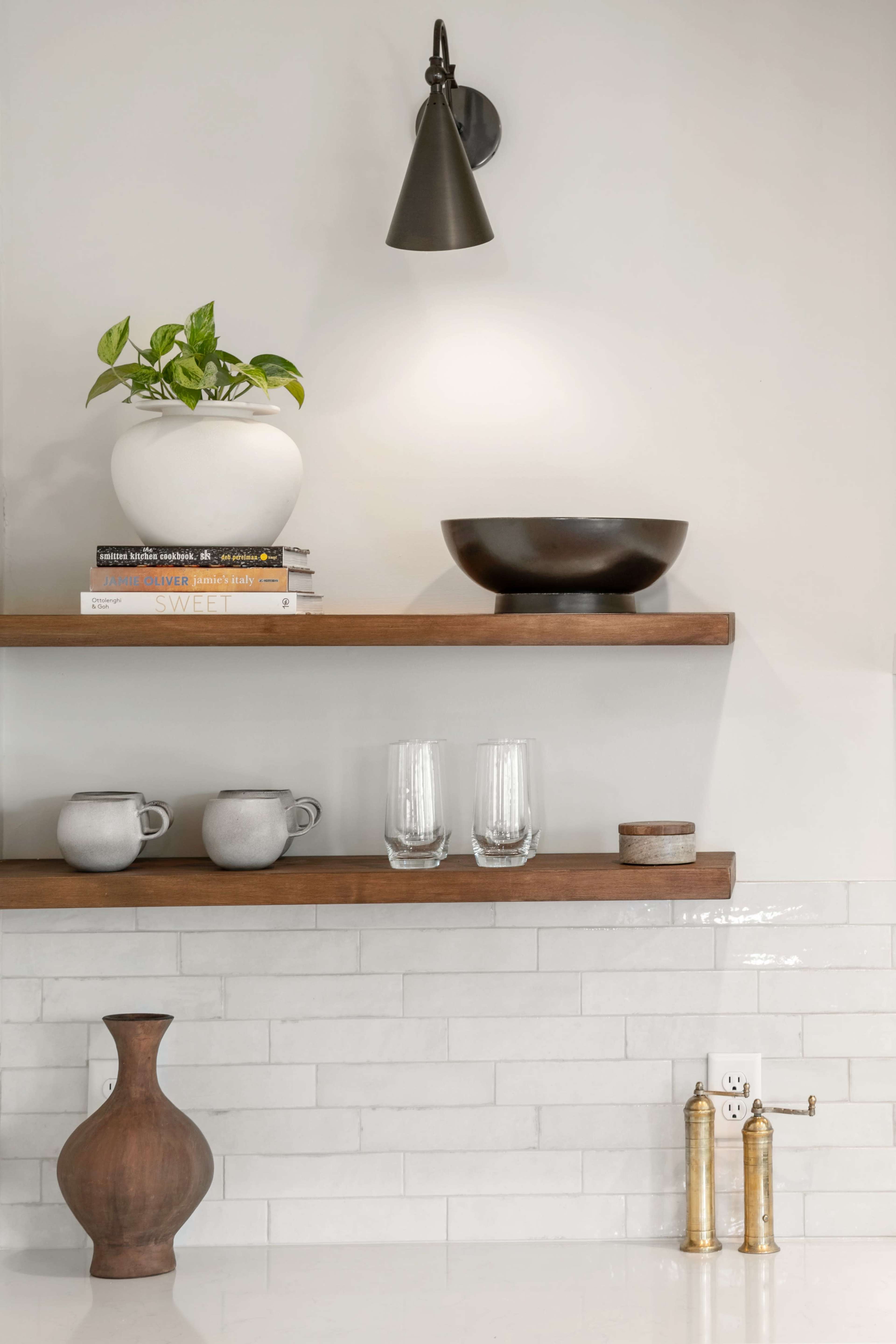 The image features a minimalist kitchen shelf with a planter, books, bowls, glasses, and a vase, complemented by a wall-mounted light fixture and a faucet below.