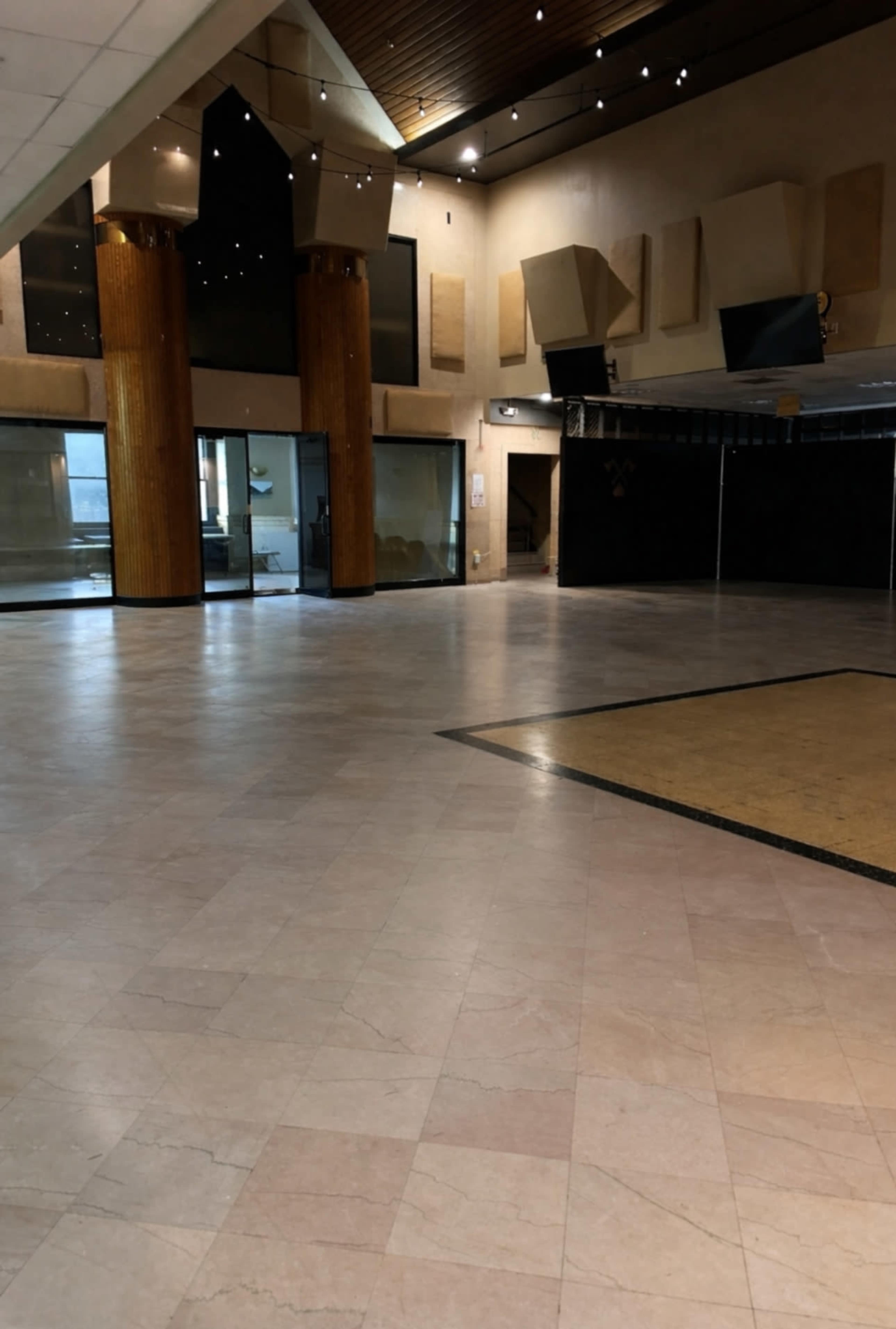 The image shows a spacious, empty indoor area with large windows, polished stone floors, and wooden columns.
