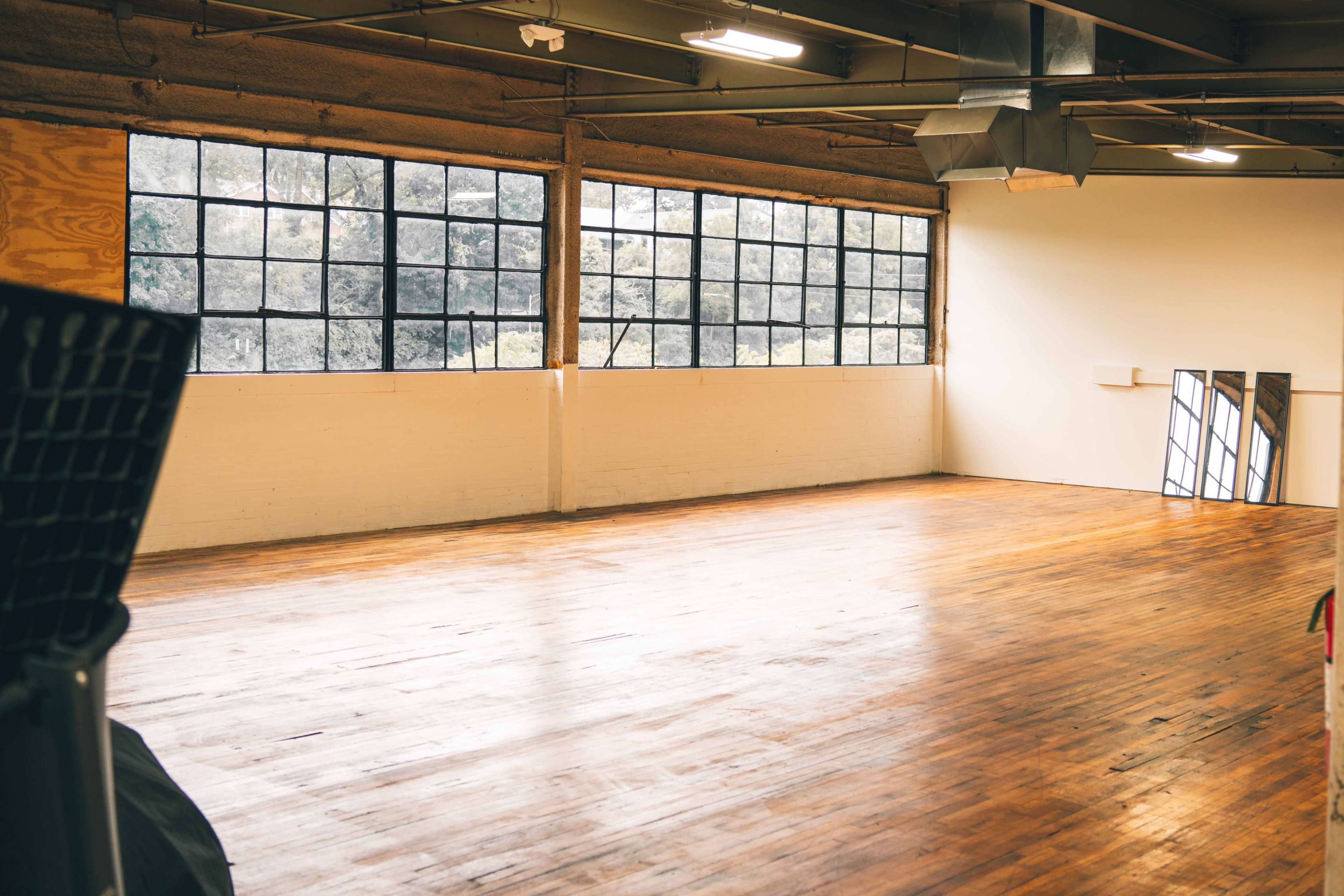 The image shows a spacious room with polished wooden flooring, large windows, and minimal furnishings.