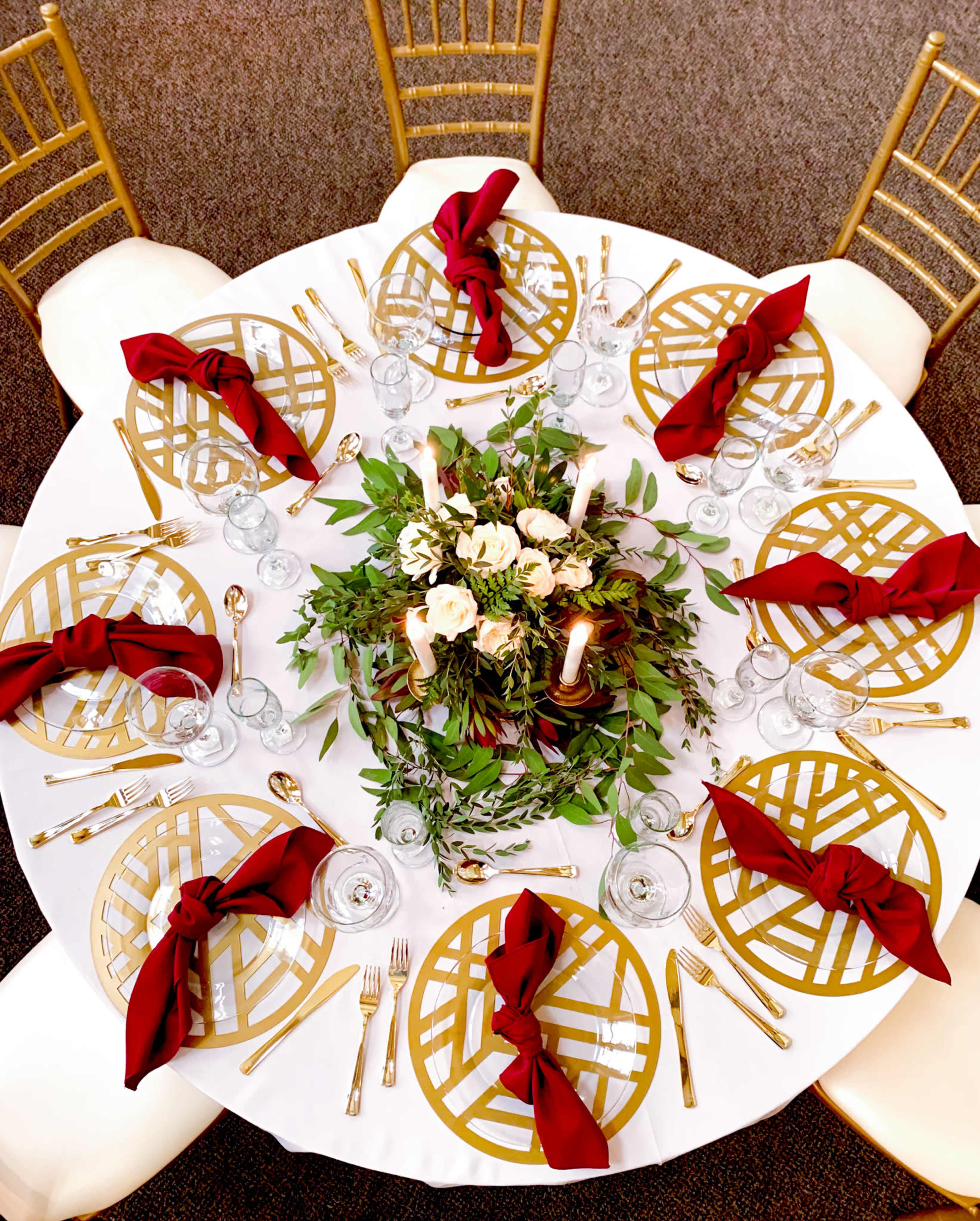 A round dining table is set with gold-patterned placemats, red napkins, glassware, and a floral centerpiece surrounded by candles.