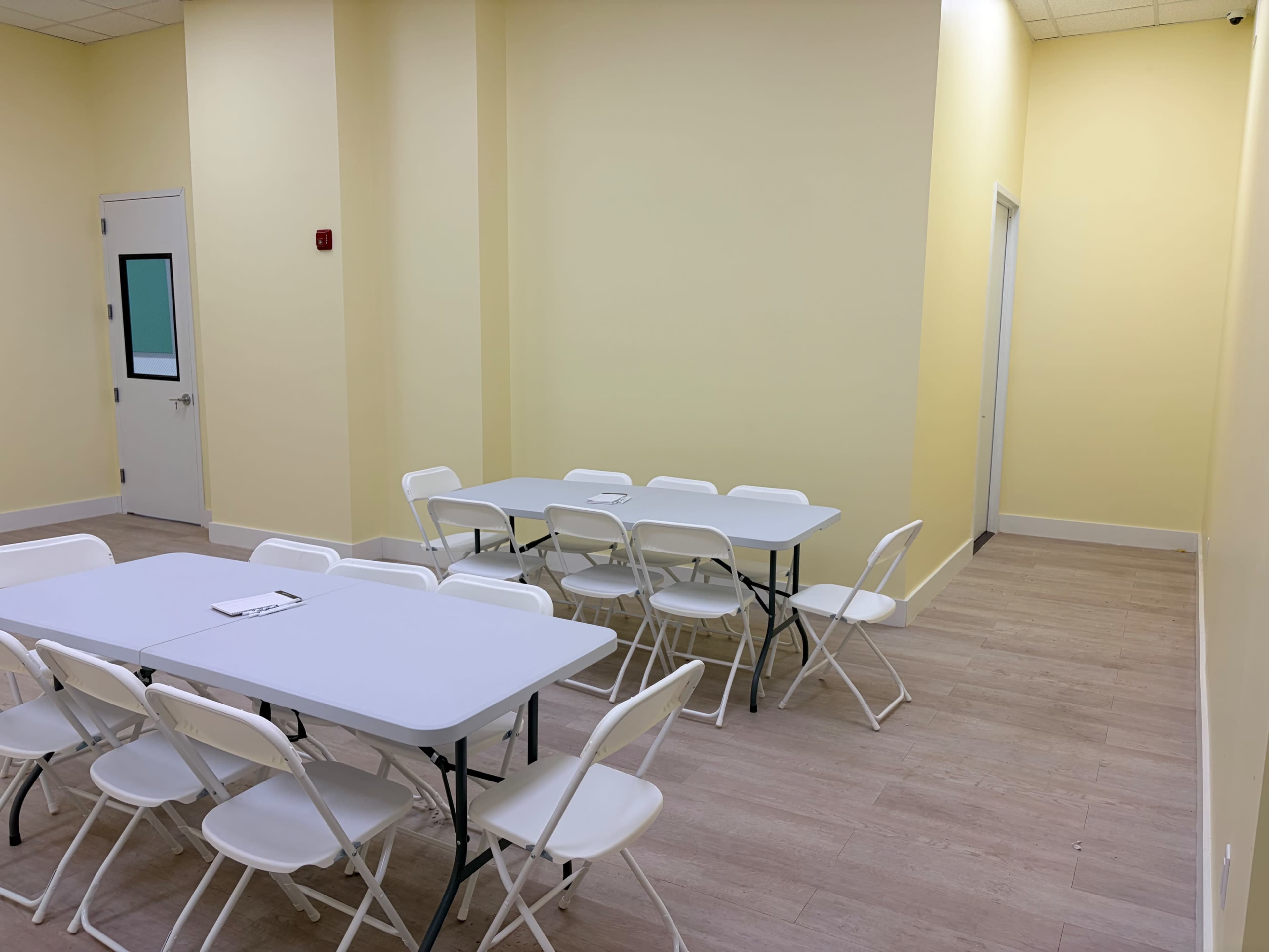 The image shows a light yellow room with two rows of folding tables surrounded by chairs on a wooden floor.