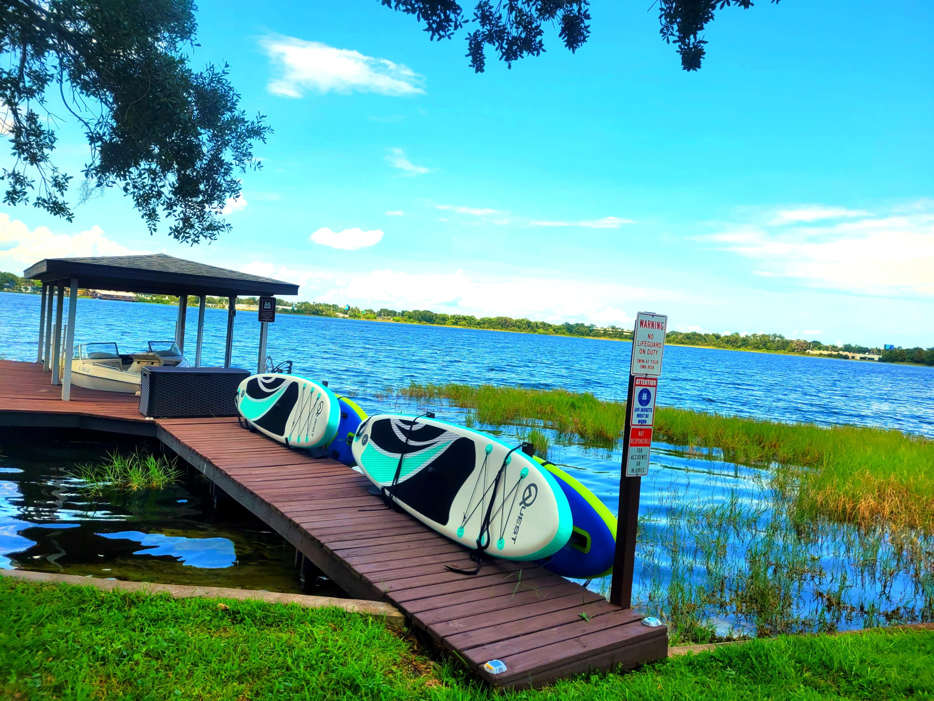 Two paddleboards are resting on a wooden dock by a lake, with grassy shoreline and a gazebo in the background.