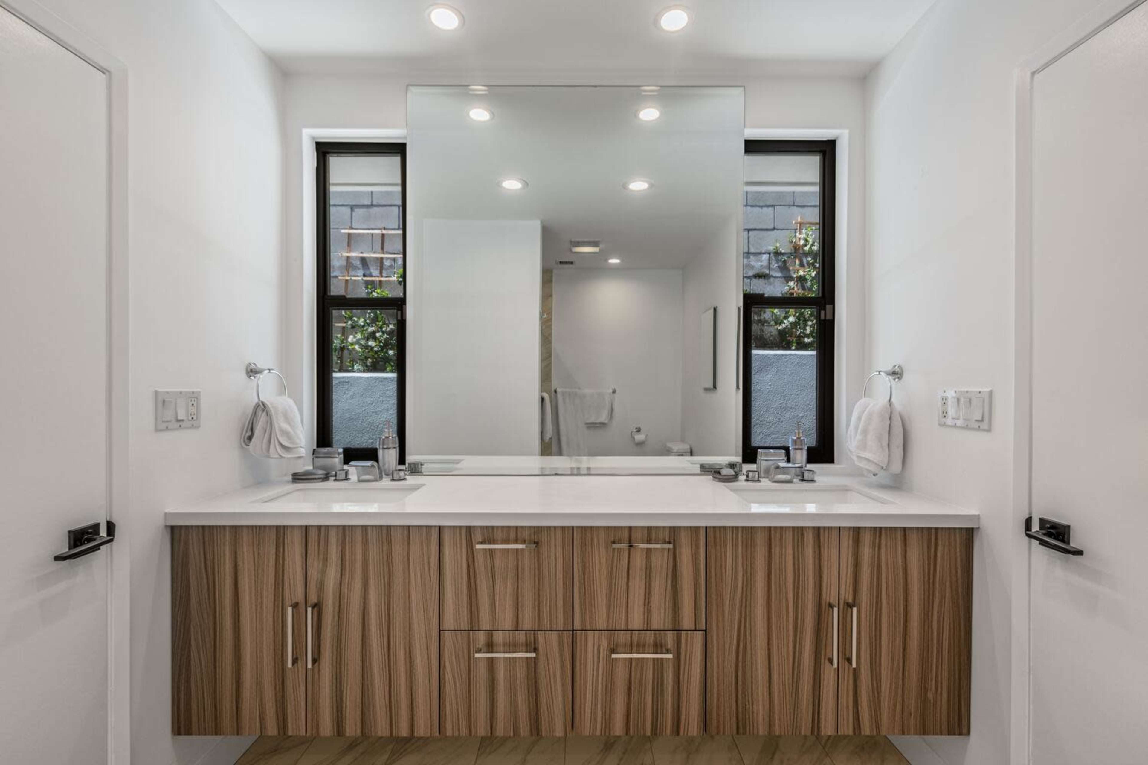 A modern bathroom with a double vanity, large mirror, and windows on either side allowing natural light to enter.