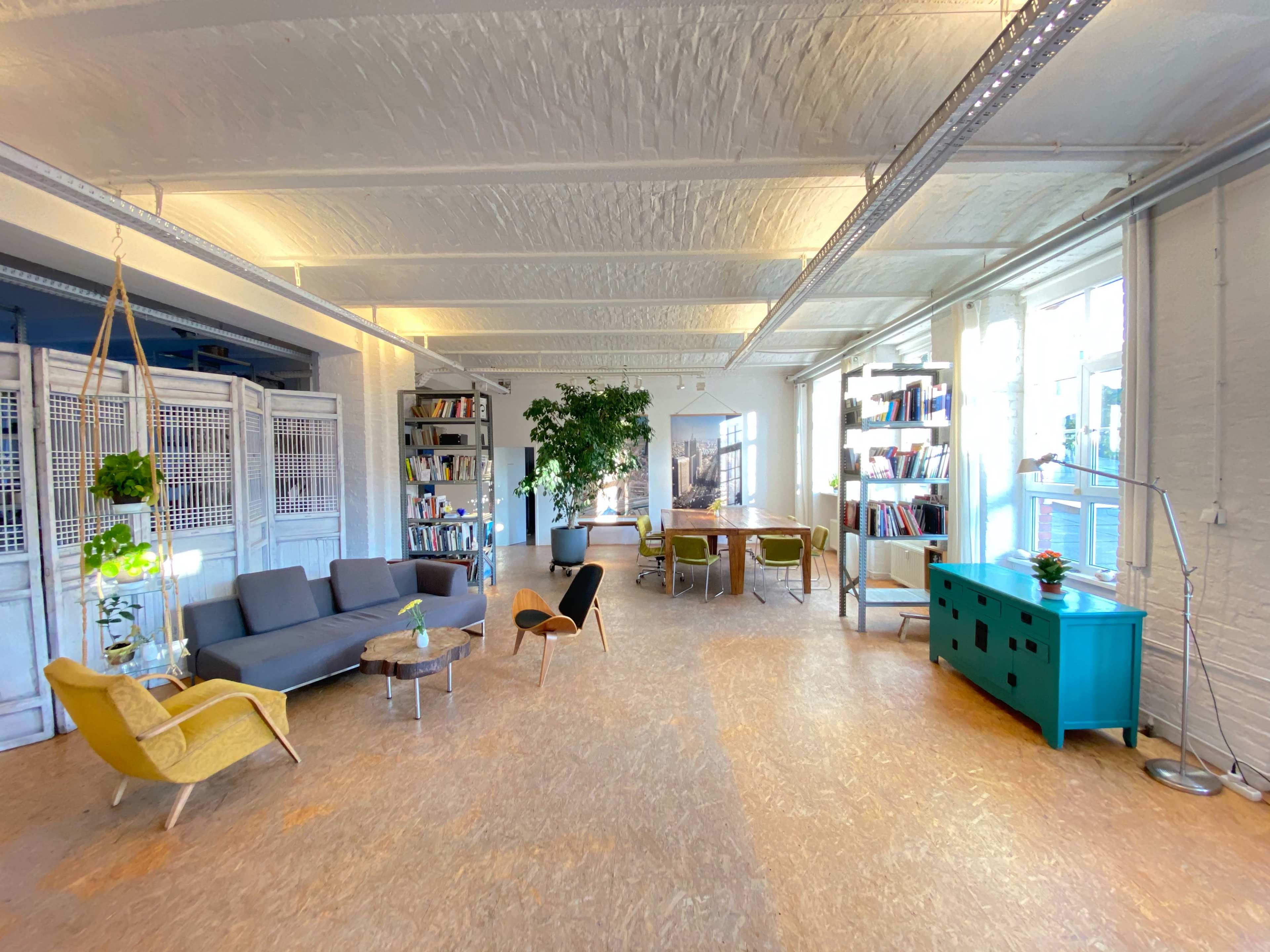 A spacious office interior with a lounge area featuring a gray sofa, yellow chair, wooden table, bookshelves, and large windows that allow natural light to enter.