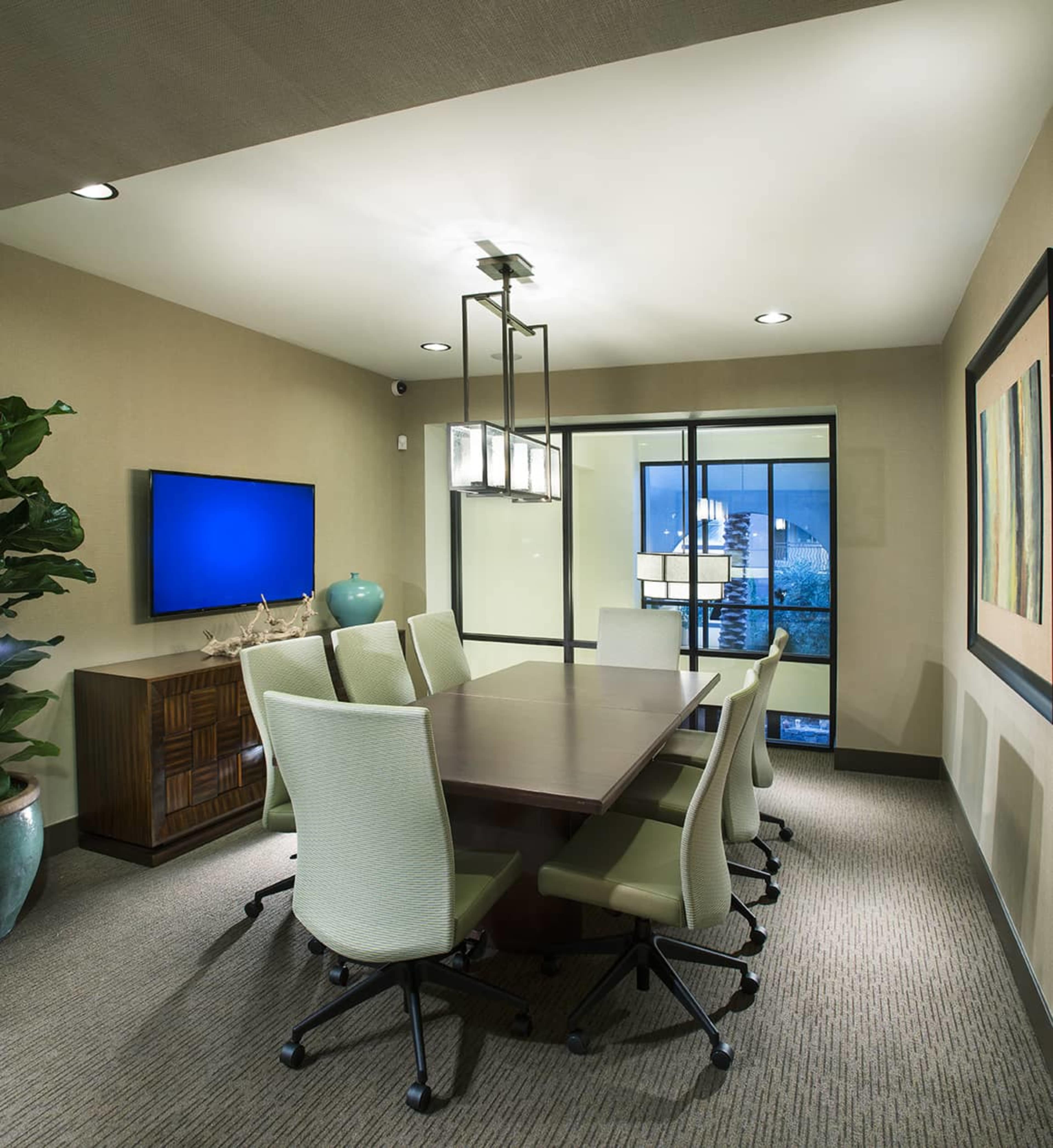 A modern conference room features a long wooden table surrounded by six office chairs, a wall-mounted TV, and large windows with an outdoor view.