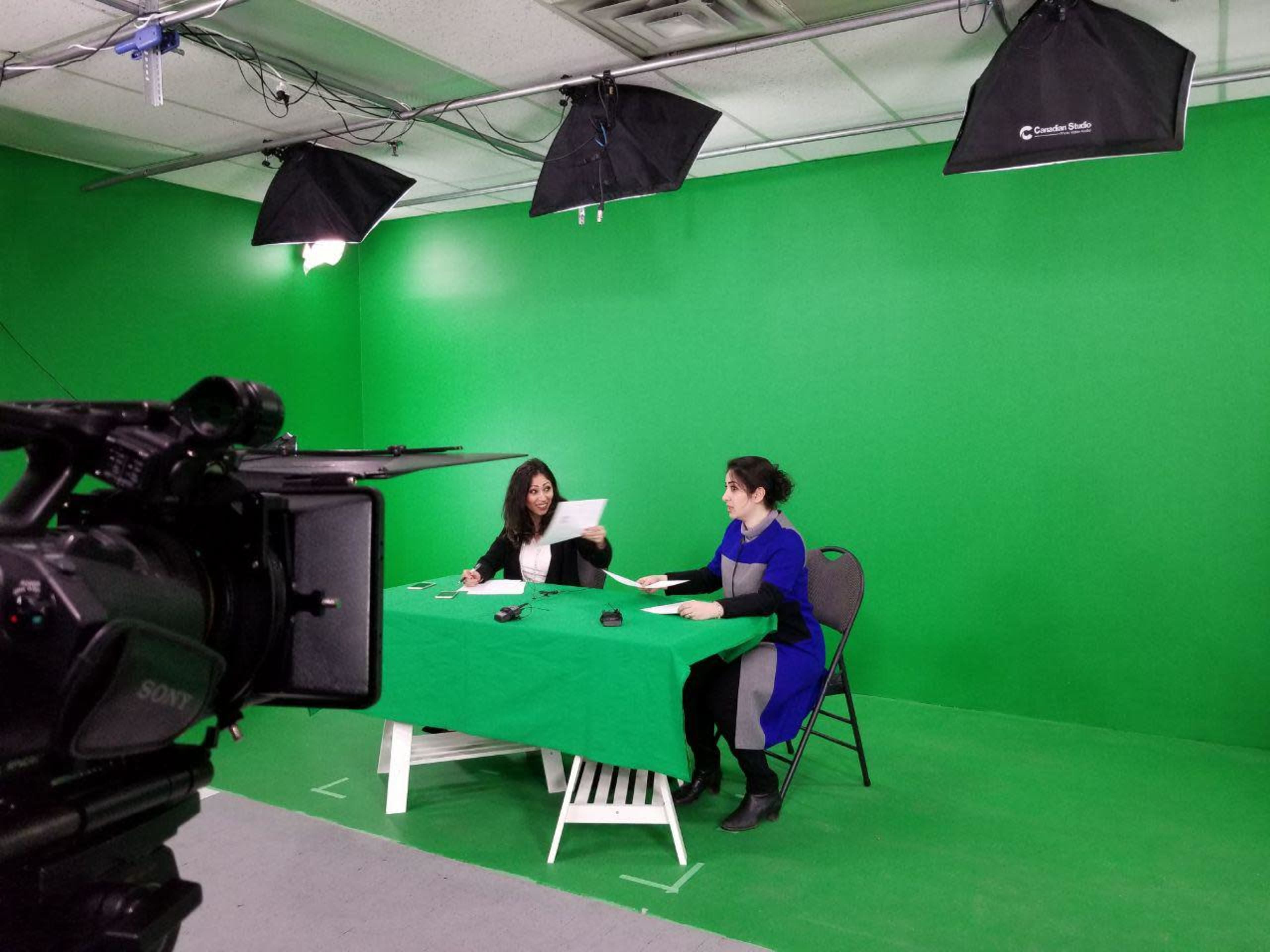 Two women sit at a table covered with a green cloth in front of a green screen, preparing for a video shoot under multiple studio lights.