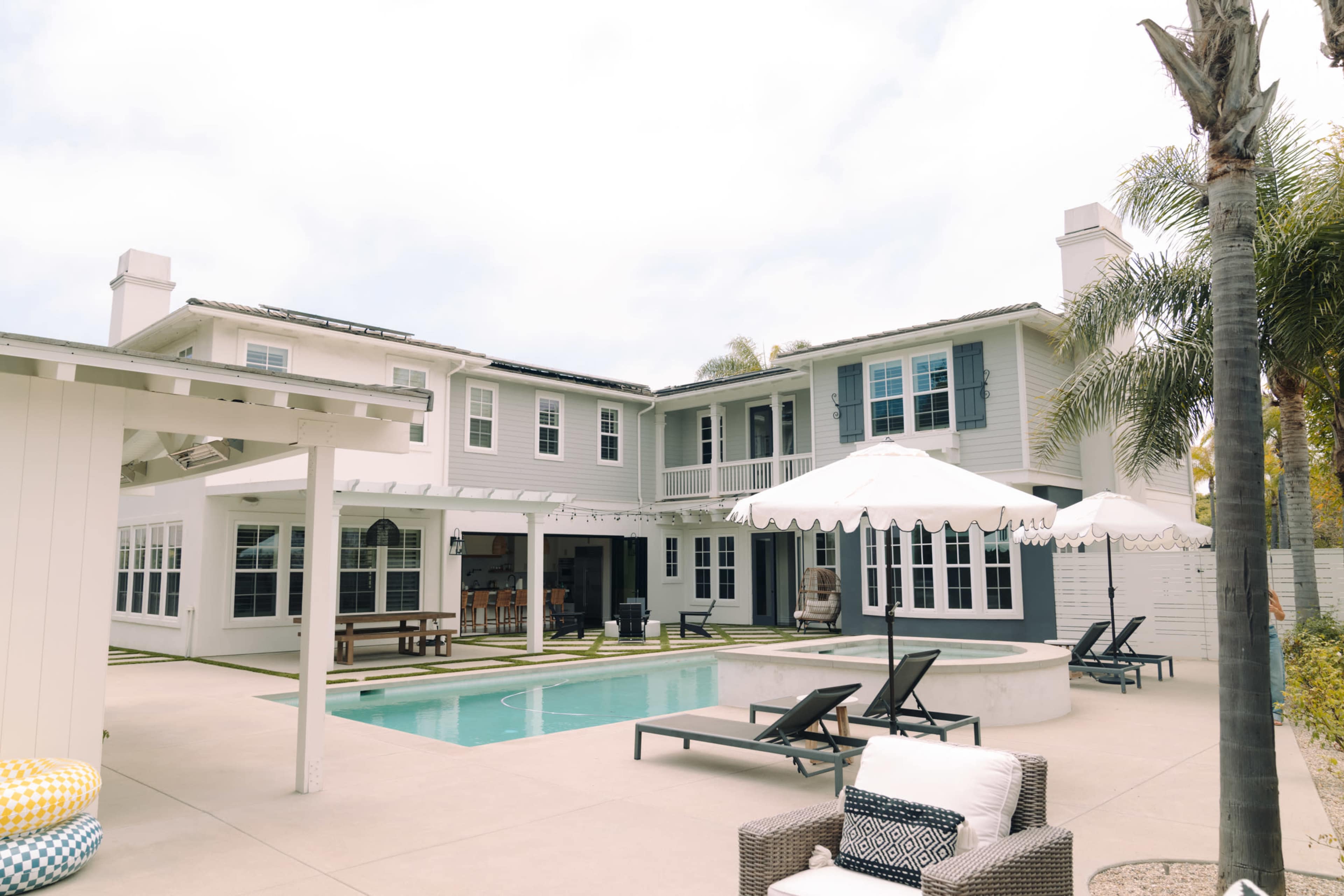 The image shows a modern home with multiple stories, a swimming pool, and patio furniture, surrounded by palm trees.