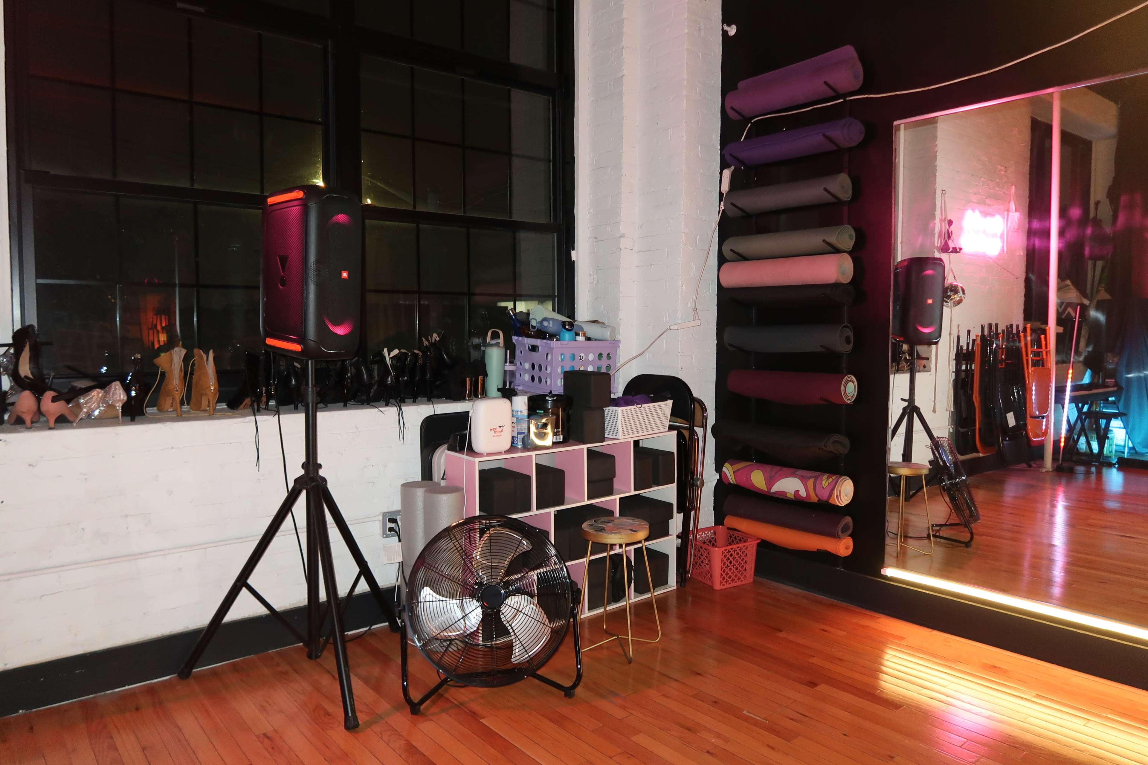 The image shows a dance studio with exercise mats stacked on a wall, a fan on the floor, and sound equipment set up near a large window.