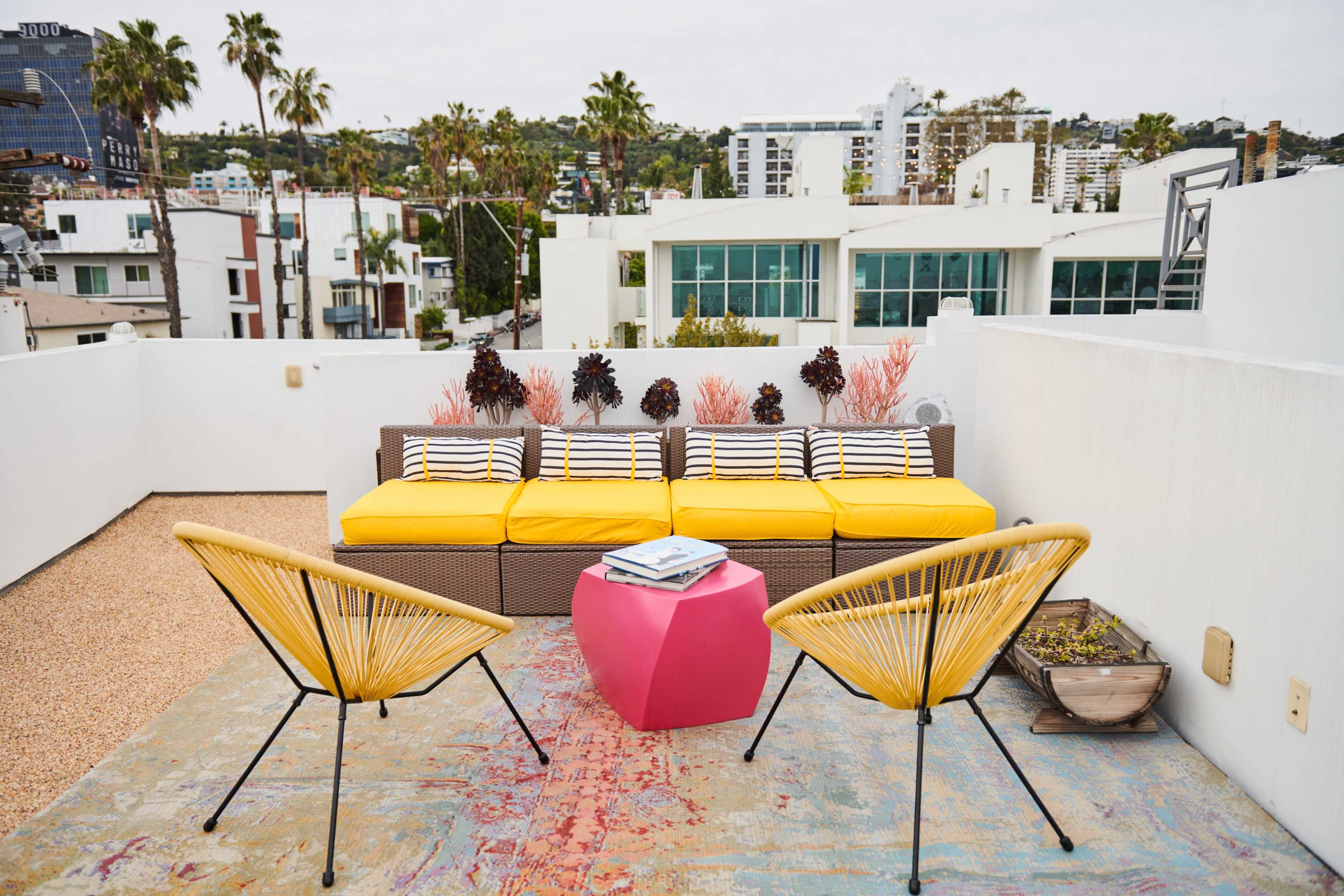A rooftop terrace featuring a yellow couch with striped cushions, two black chairs, a pink side table, and potted plants, against a backdrop of buildings and palm trees.