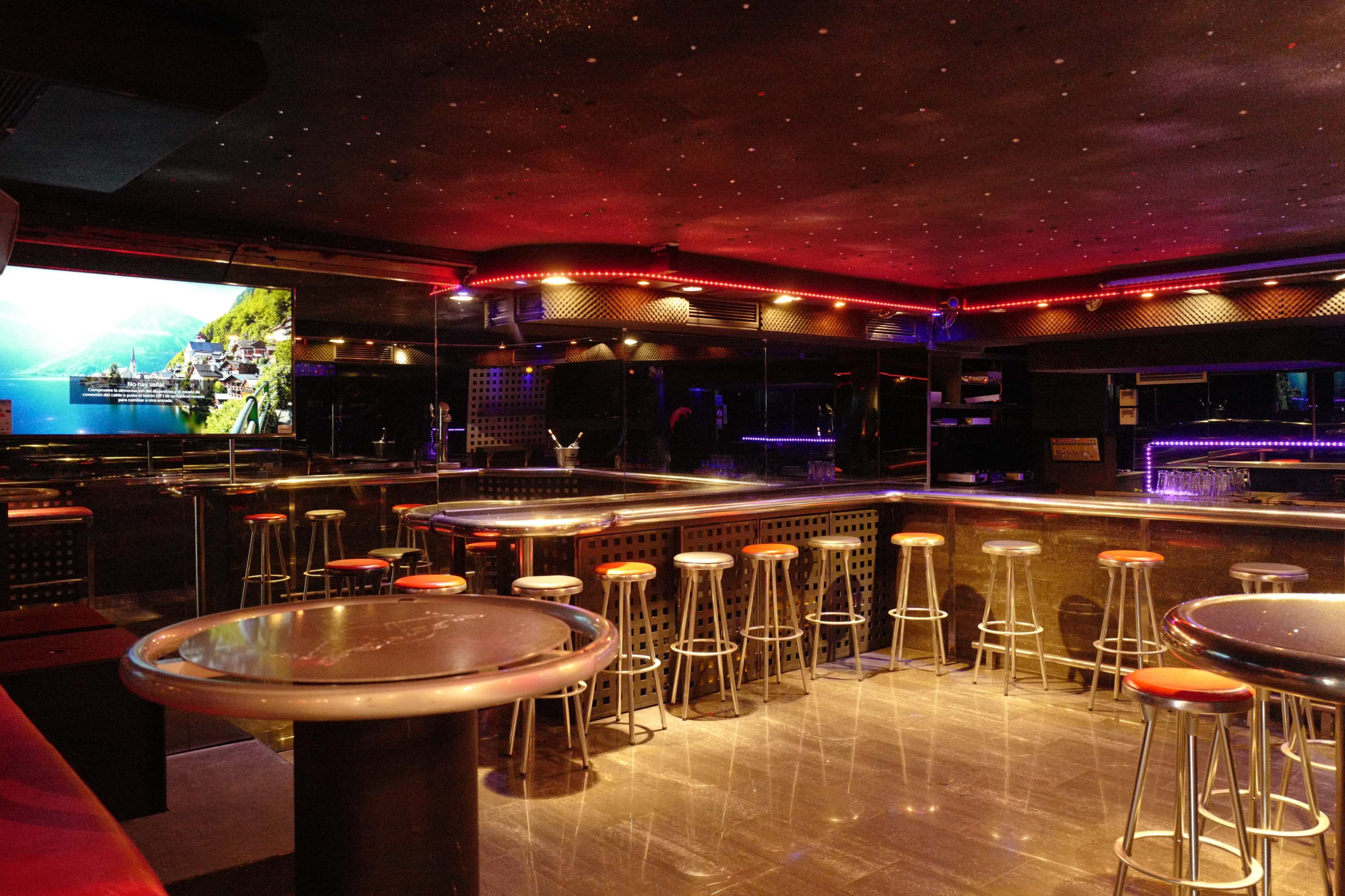 A dimly lit bar area with high stools and circular tables, featuring colorful lighting and mirrored surfaces.