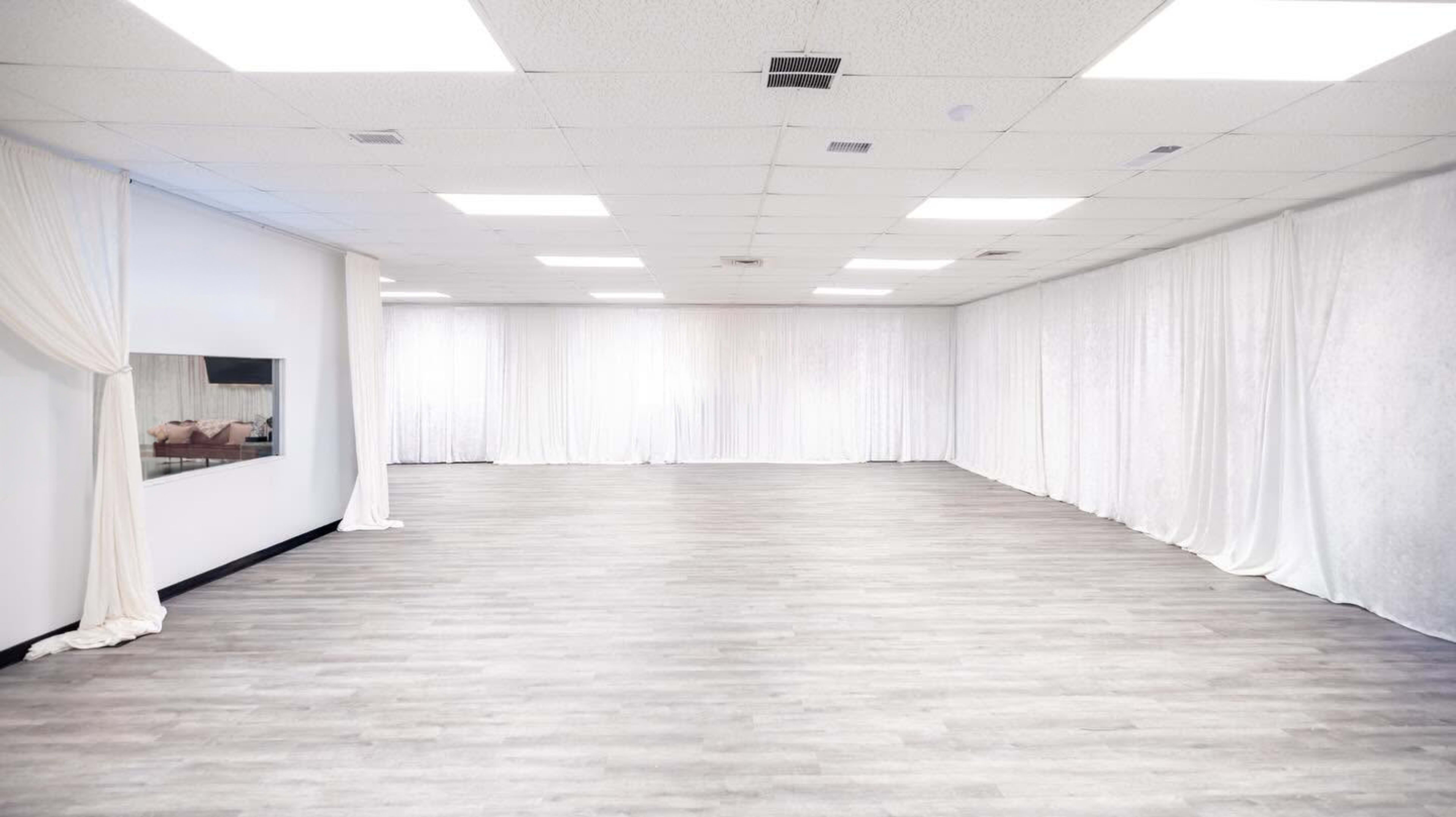 The image shows an empty room with a wooden floor, white draped curtains along the walls, and overhead fluorescent lighting.