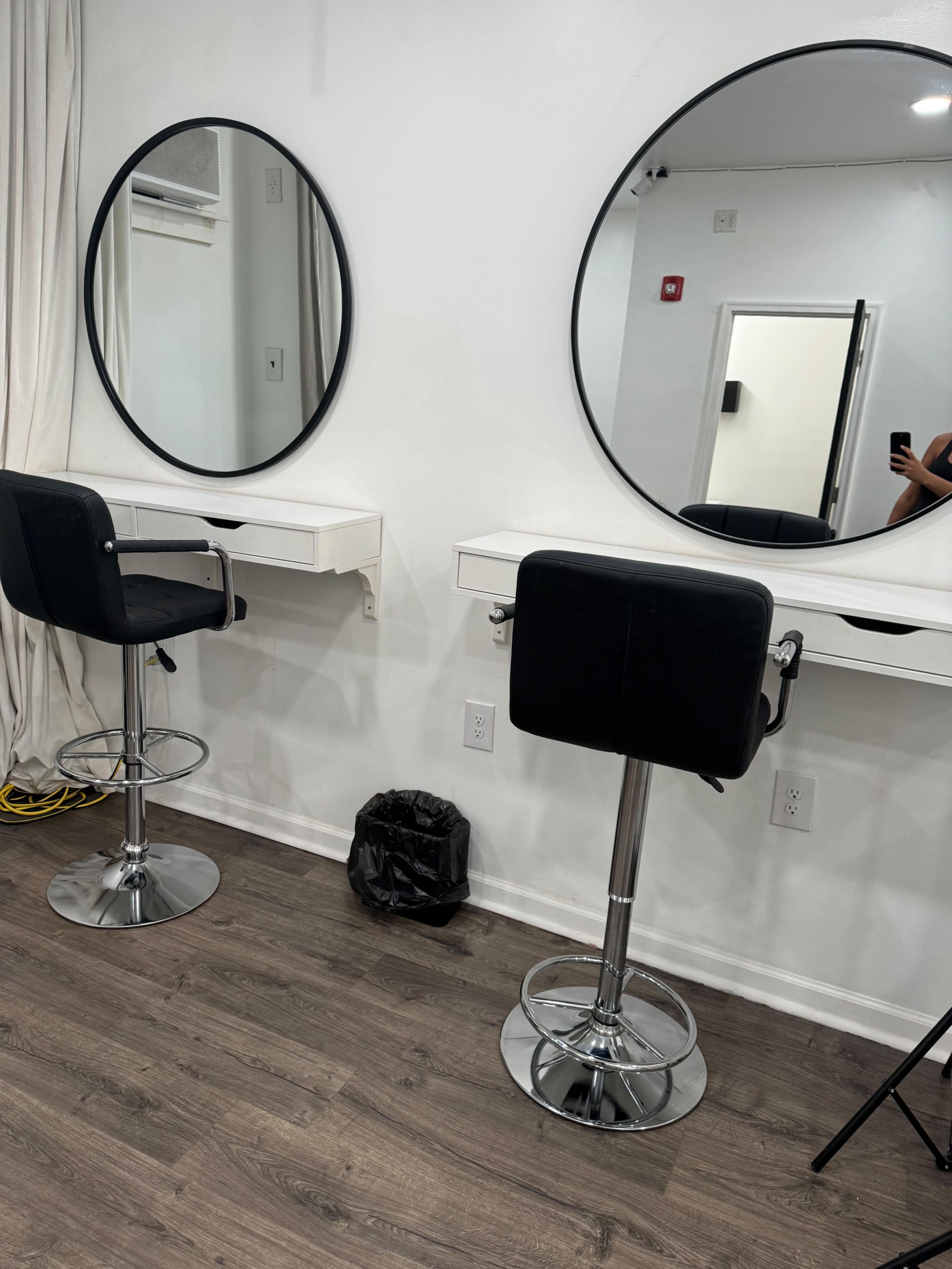 A minimalist salon interior featuring two black swivel chairs in front of circular mirrors mounted on a white wall.